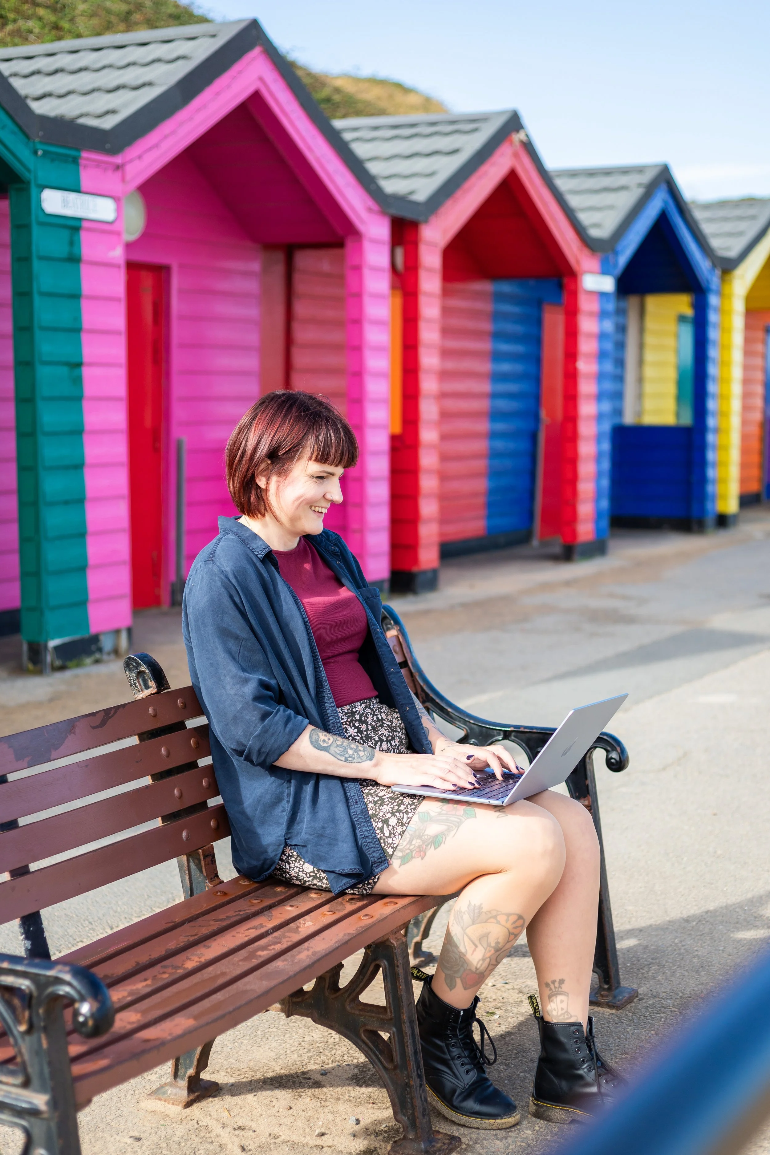 Kat with brown hair is smiling and sat on a bench on her laptop. In the background are colourful huts.