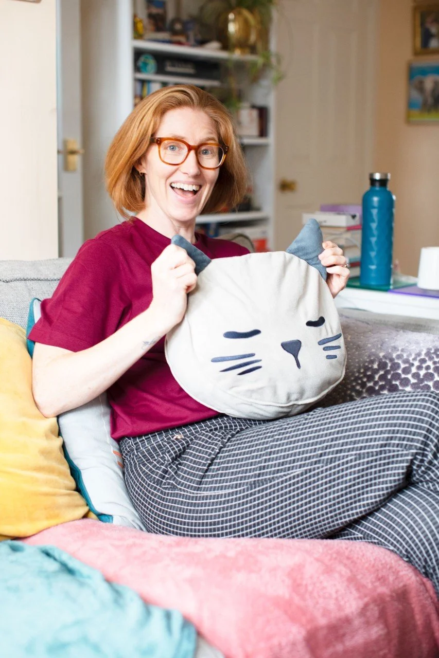 Siobhan fox holding a cat pillow and sitting on a colourful sofa. Her expression is surprised.