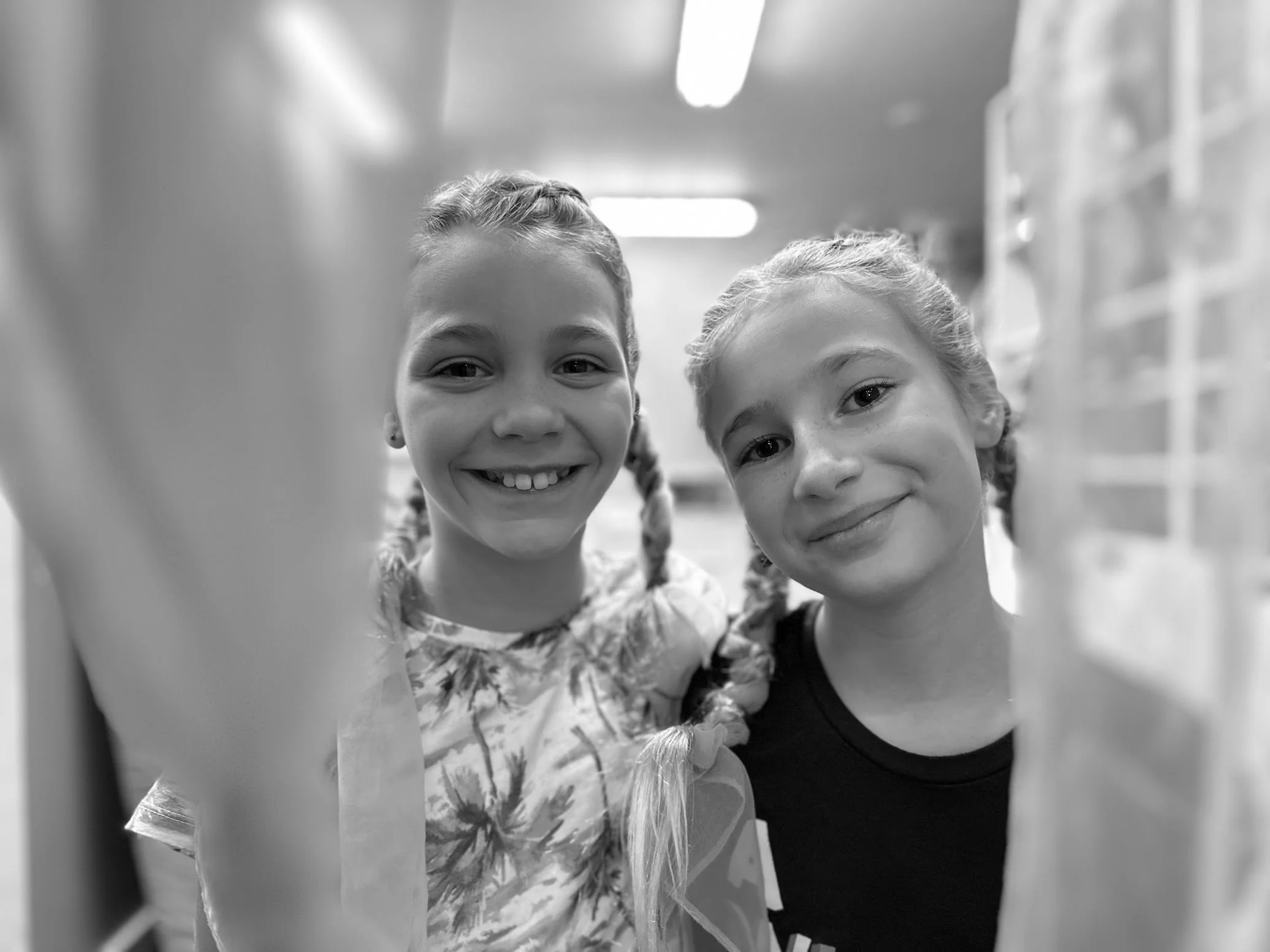 Two young girls smiling, close-up portrait in black-and-white, standing indoors with shelves in the background.