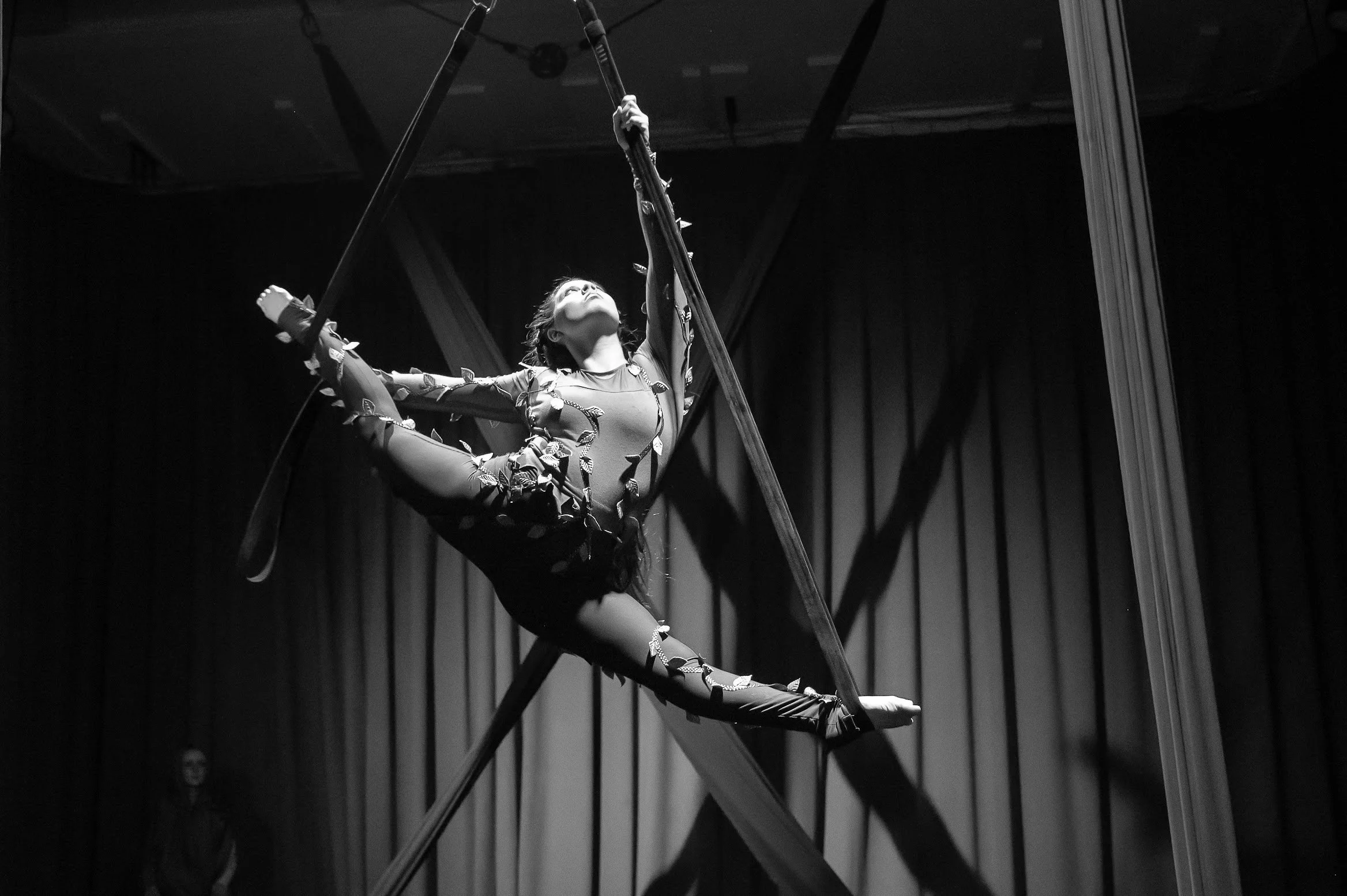 A female trapeze artist performs an aerial act on a trapeze in a darkened theater, with curtains and stage equipment in the background.