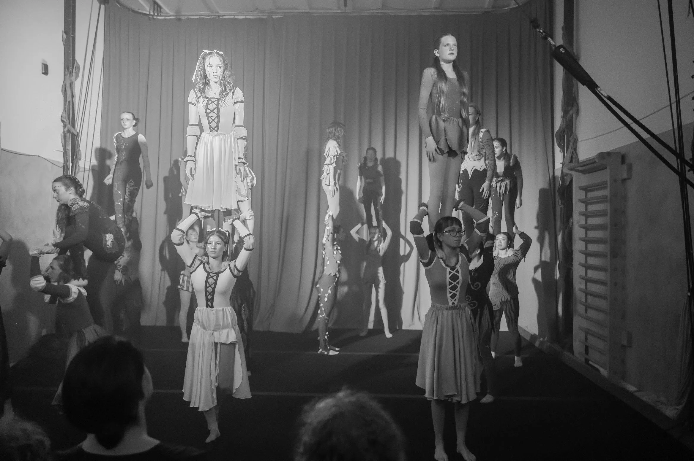 Ensemble troupe performing a circus act on stage, with some balancing on each other's shoulders and others standing or sitting in various poses. The stage has a curtain backdrop and stage equipment visible to the side.