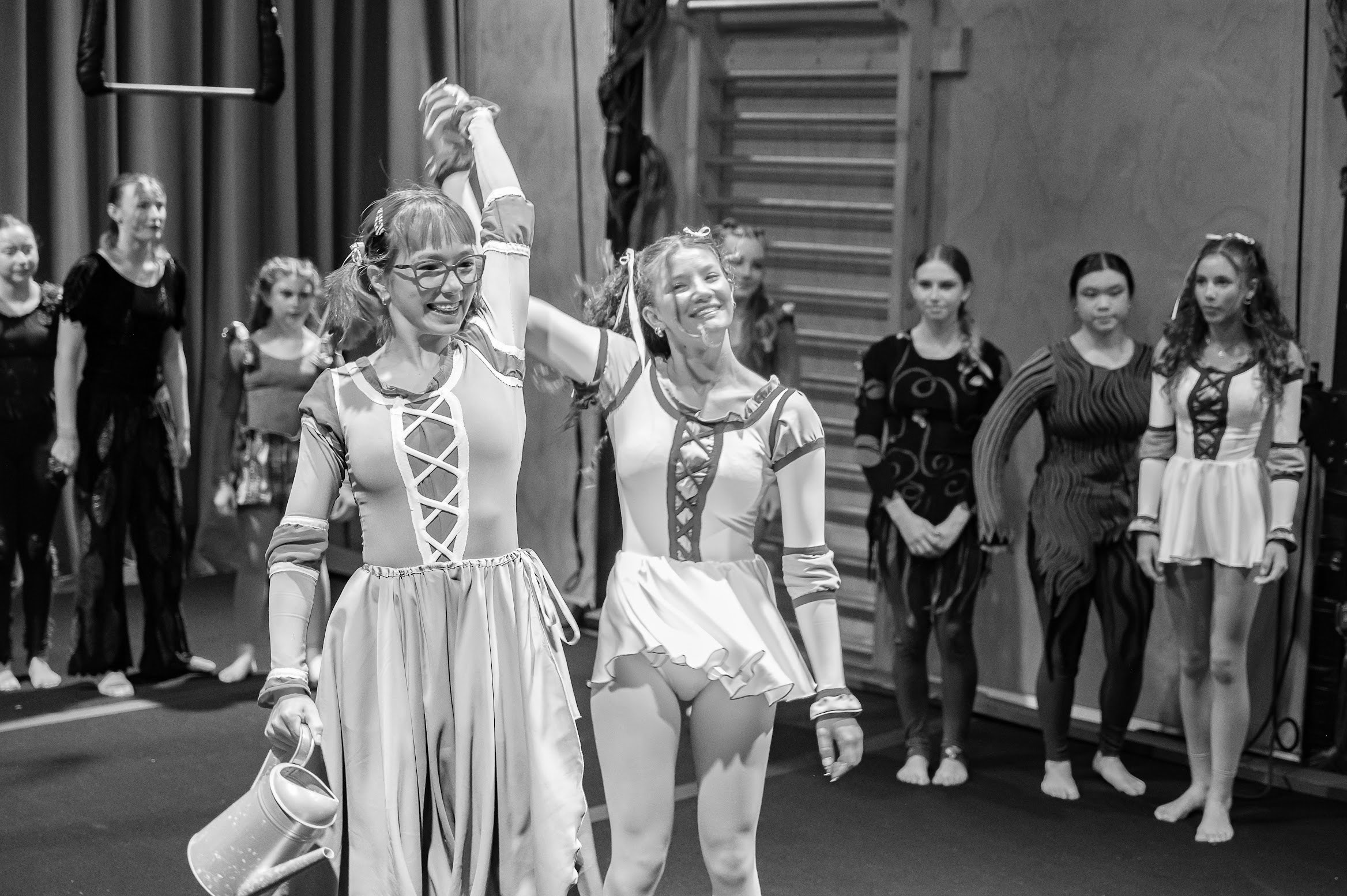 A group of people in a dance rehearsal in a gymnasium. The women in the foreground are smiling and holding ballet shoes, while the girls in the background are dressed in costumes, standing in front of a backdrop with gym equipment.