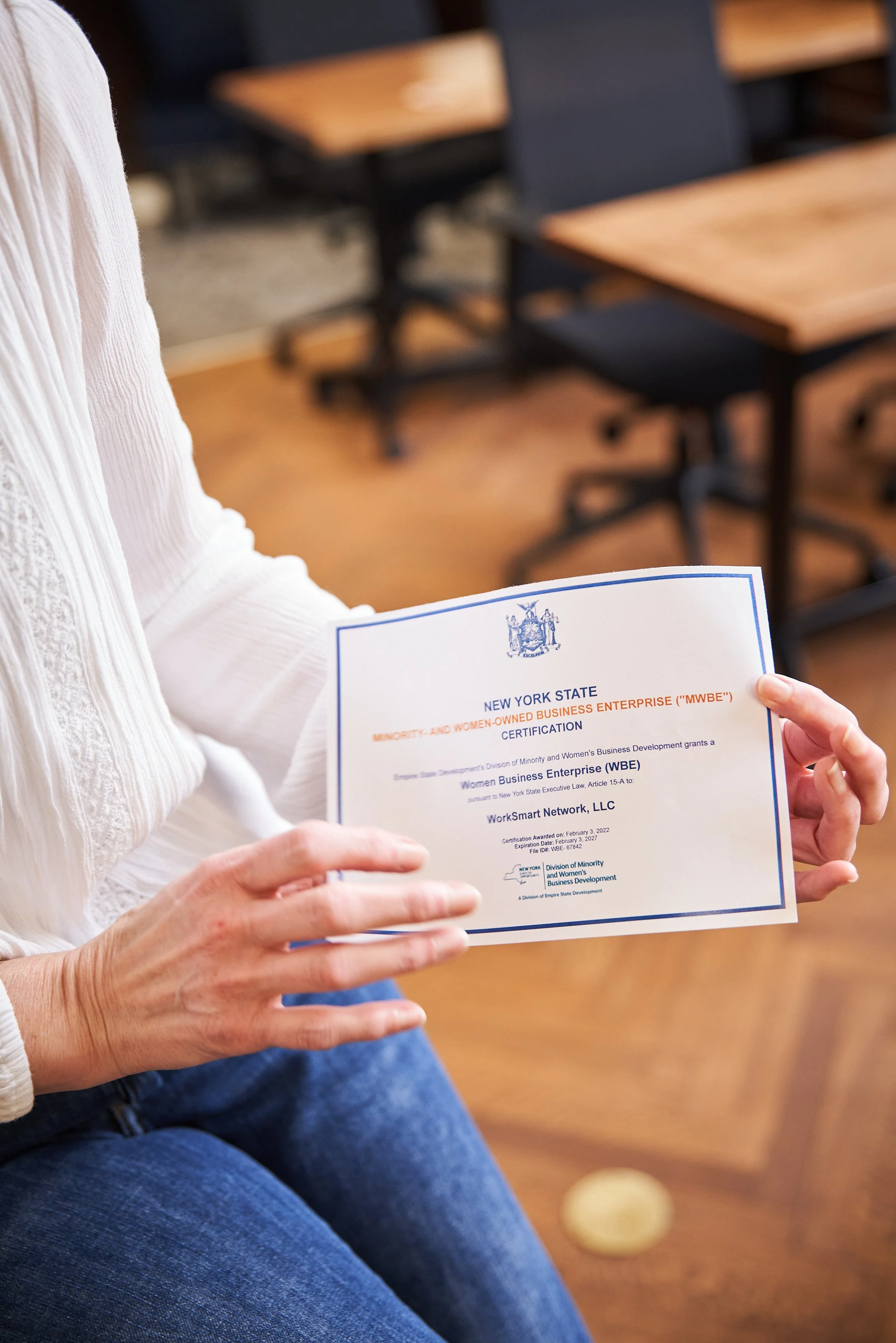 Person holding a certificate from New York State recognizing Women Business Enterprise (WBE) certification, seated in a room with wooden floors and black chairs.