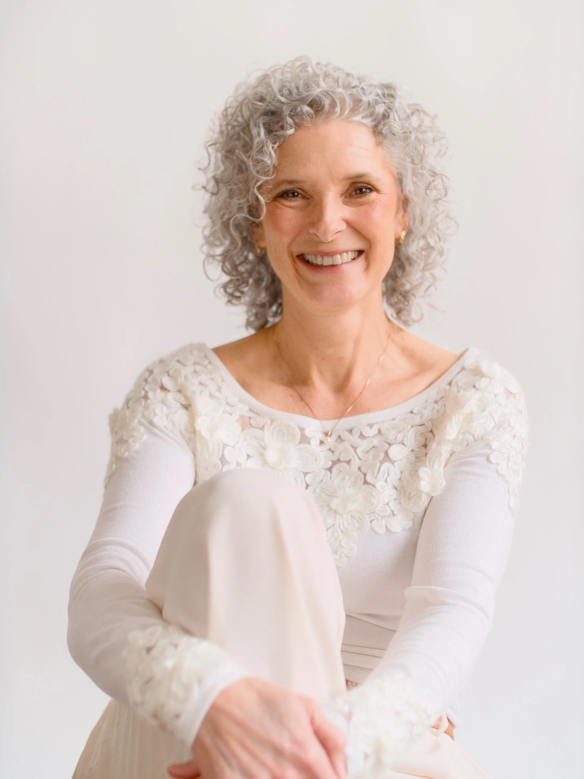 A woman with short curly gray hair sitting against a plain white background, smiling, wearing an elegant cream-colored dress with floral embroidery on the sleeves and shoulders.