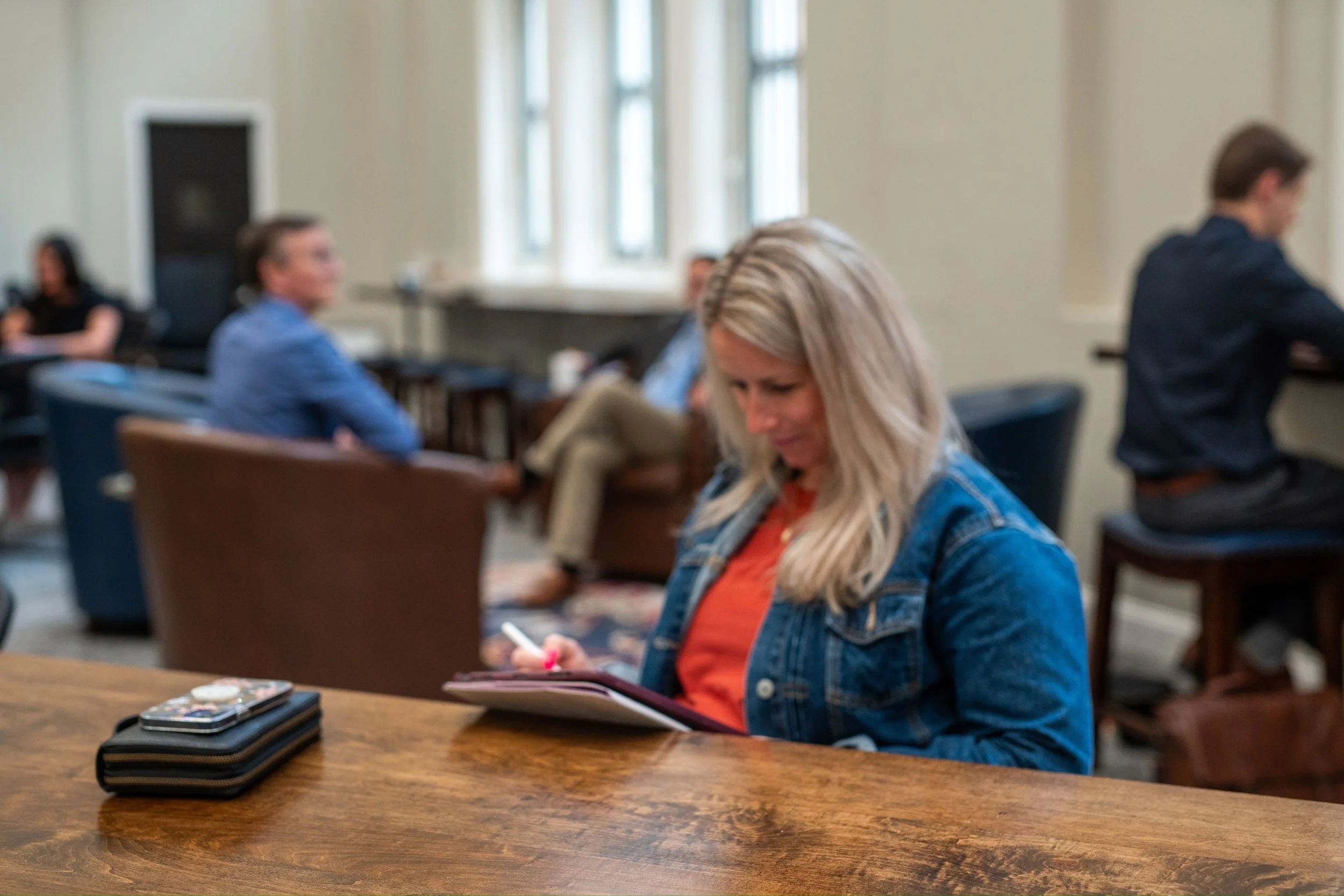 A woman in the foreground is working on her technology. In the background furniture and other men and women are working and talking in a coworking space.
