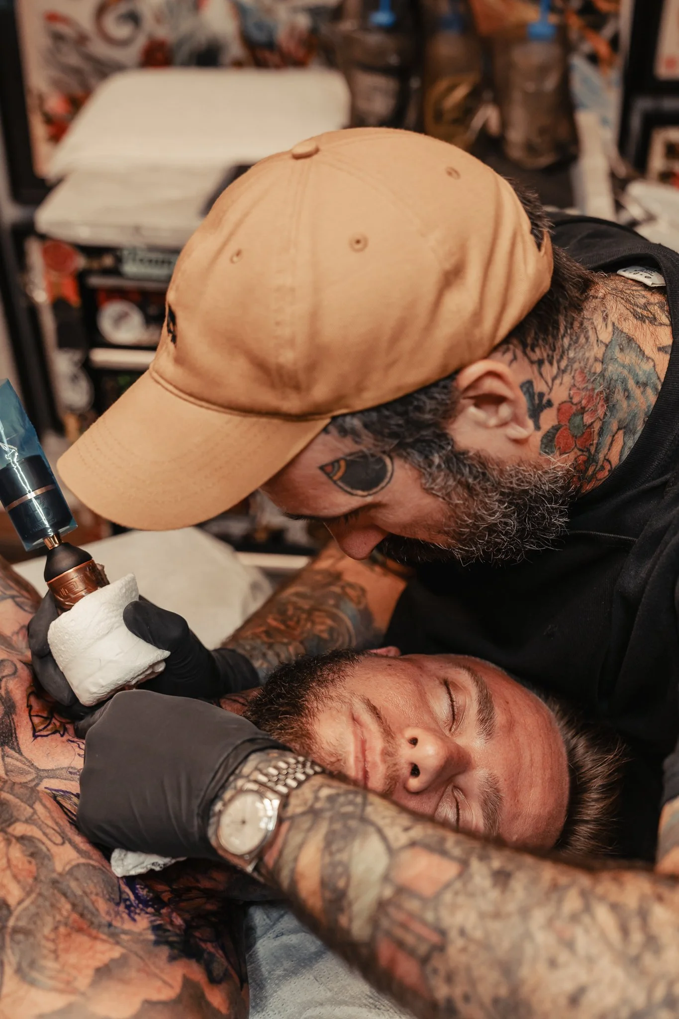 A tattoo artist is tattooing a man's arm while he is lying down with his eyes closed. The artist is wearing a tan cap, black gloves, and has tattoos on his neck and arms. The background shows shelves with various supplies.