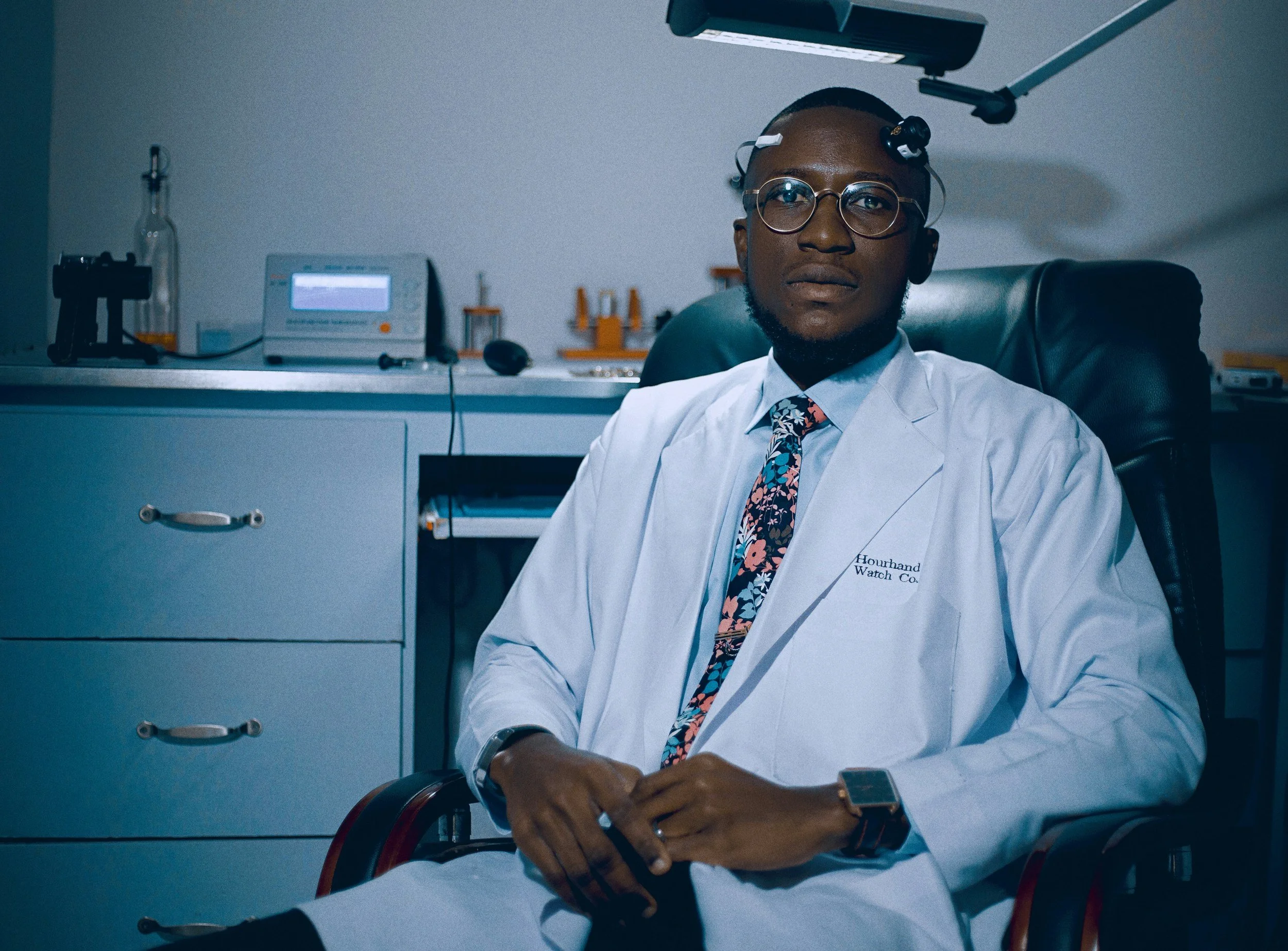 A young man wearing glasses, a white lab coat, and a floral tie, sitting in a black office chair in a medical or laboratory setting with medical equipment and supplies in the background.