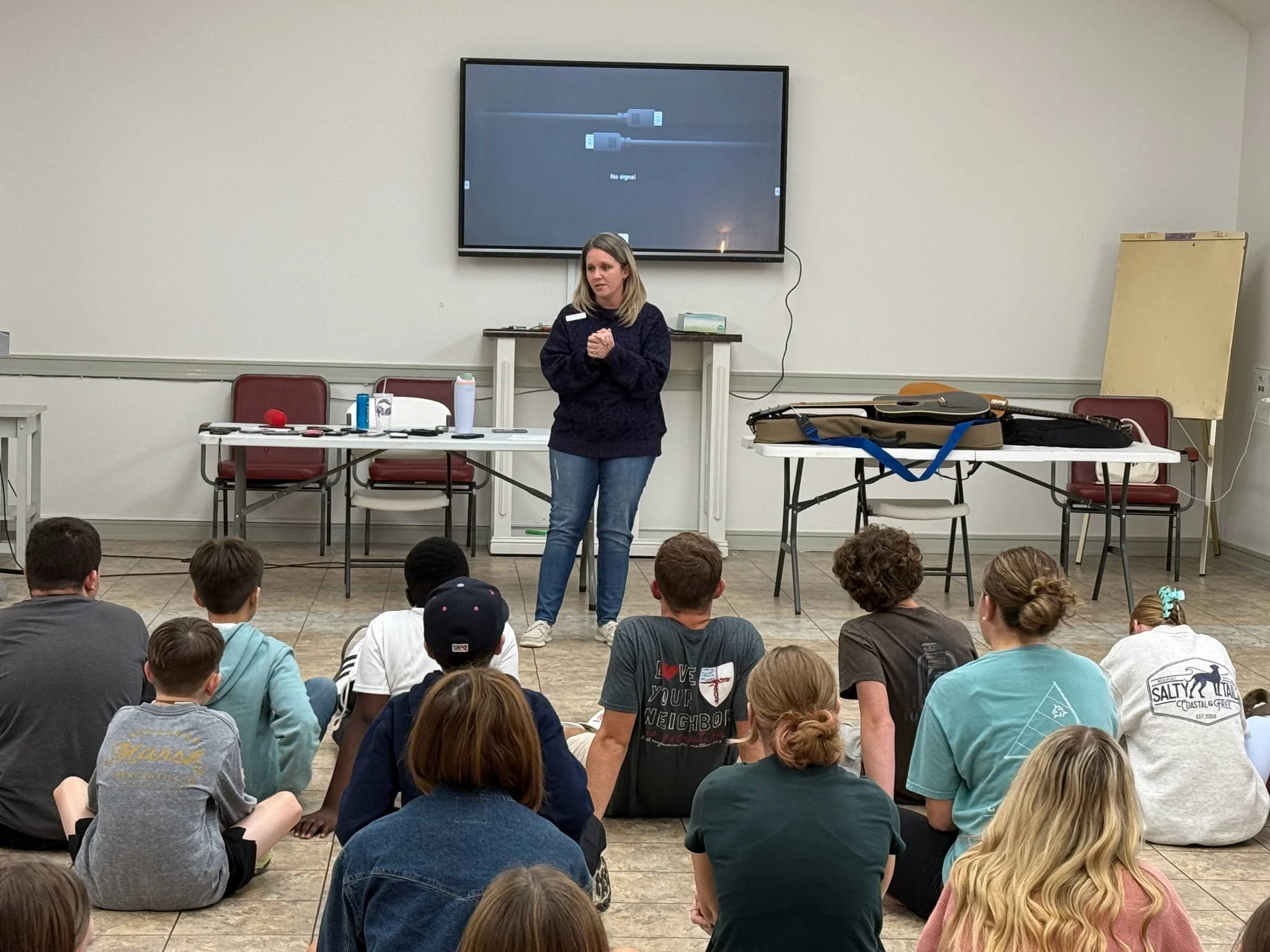 A woman stands in front of a group of children seated on the floor, giving a presentation. A large screen behind her displays a no signal message. Tables with supplies are on either side of her.