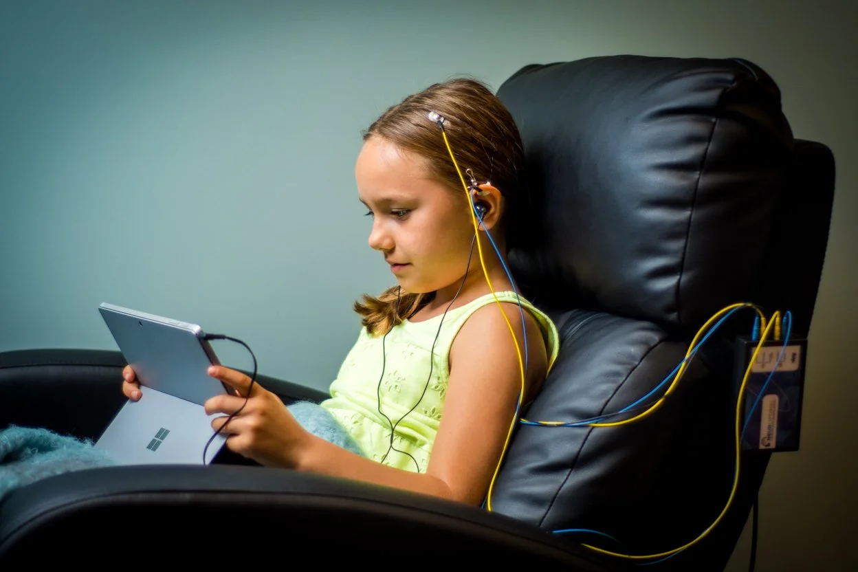 A young girl sitting in a black reclining chair, looking at a handheld gaming device or tablet, receiving NeurOptimal® neurofeedback, with a neutral-colored wall in the background.