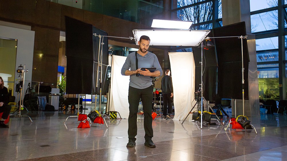 Man standing in front of a photo studio setup in a modern indoor space, looking at a tablet, with lighting equipment and backdrop screens around him.