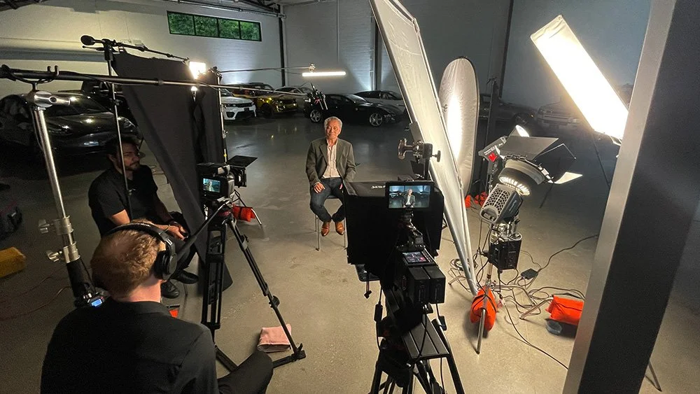 A man sitting on a stool being filmed in a professional studio with cameras, lighting, and equipment, inside a garage with cars in the background.
