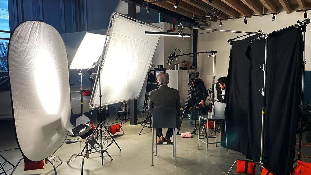 A video or photo shoot setup with professional lighting, cameras, and black curtains, with a person sitting on a chair in the center, facing away from the camera.