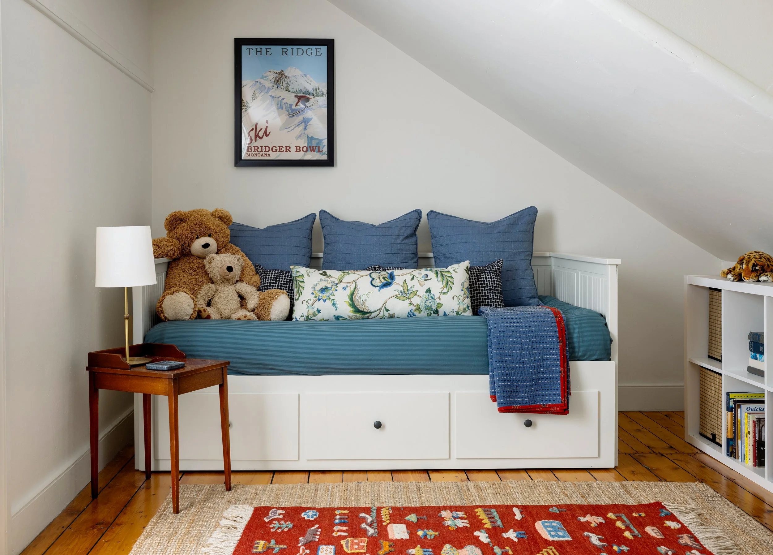A cozy bedroom corner with a white daybed featuring multiple blue pillows, a floral throw pillow, and plush teddy bears. A small wooden side table with a white lamp stands next to the bed. A framed vintage skiing poster hangs on the wall above the be