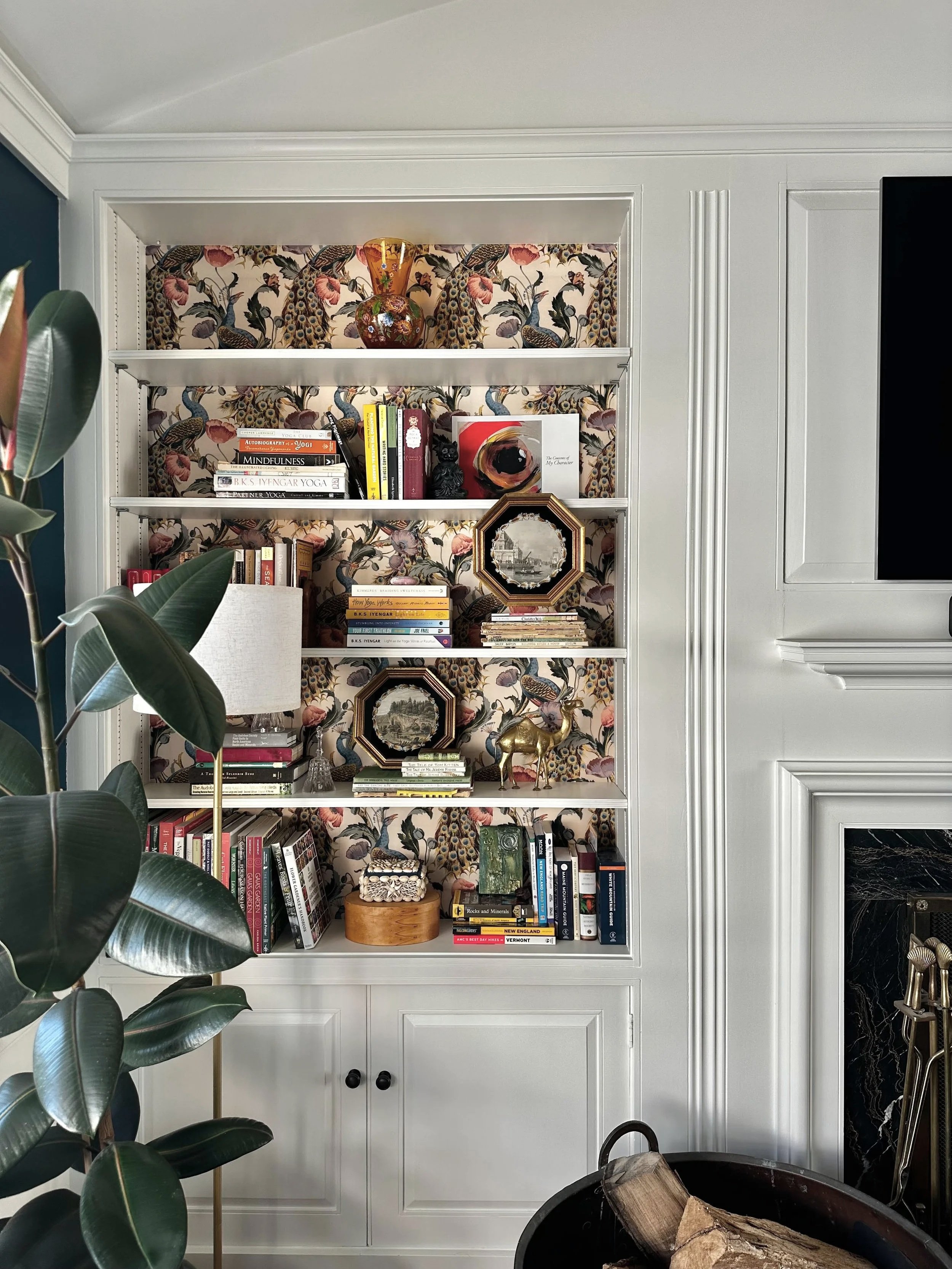 A built-in white bookshelf with decorative items, books, and framed artwork, set against floral wallpaper with peacock design. To the right is a black fireplace, and there's a large houseplant in the foreground.