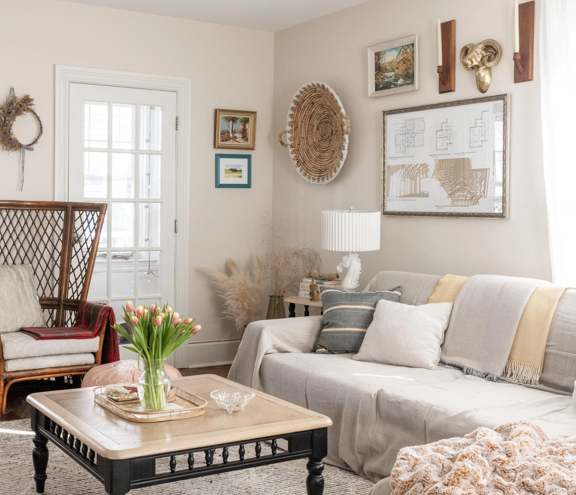 Living room with a beige sofa, pillows, a wooden coffee table with a vase of pink tulips, artwork, wall decor, a lamp, and a closed glass door letting in natural light.