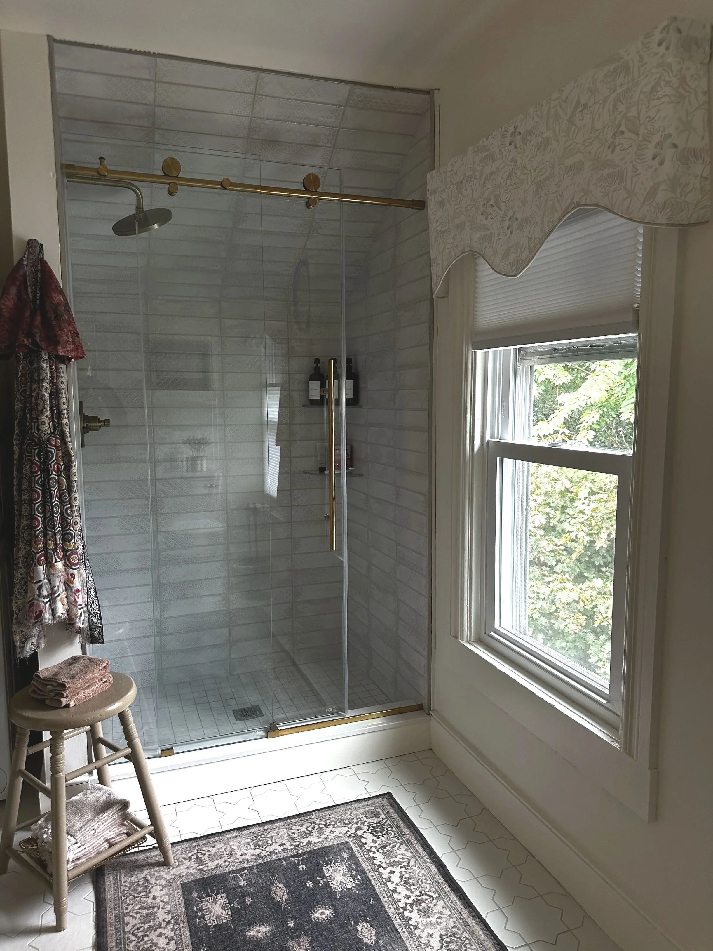 A bathroom with a glass-enclosed shower, window with blinds, and a patterned rug on the floor.