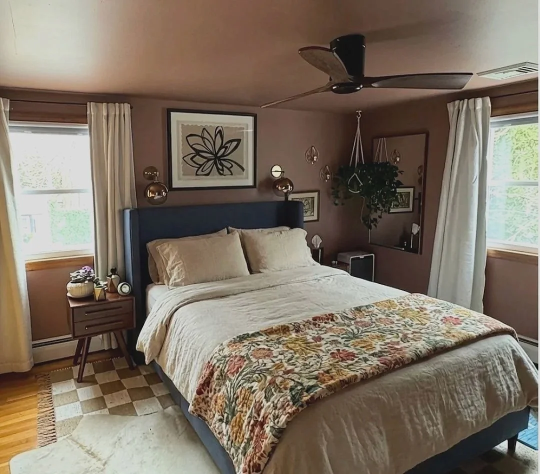 A cozy bedroom with a large bed featuring cream-colored bedding and a floral quilt. The room has two windows with white curtains, a dark blue upholstered headboard, and wall art including a large black flower sketch and framed pictures. There are bed