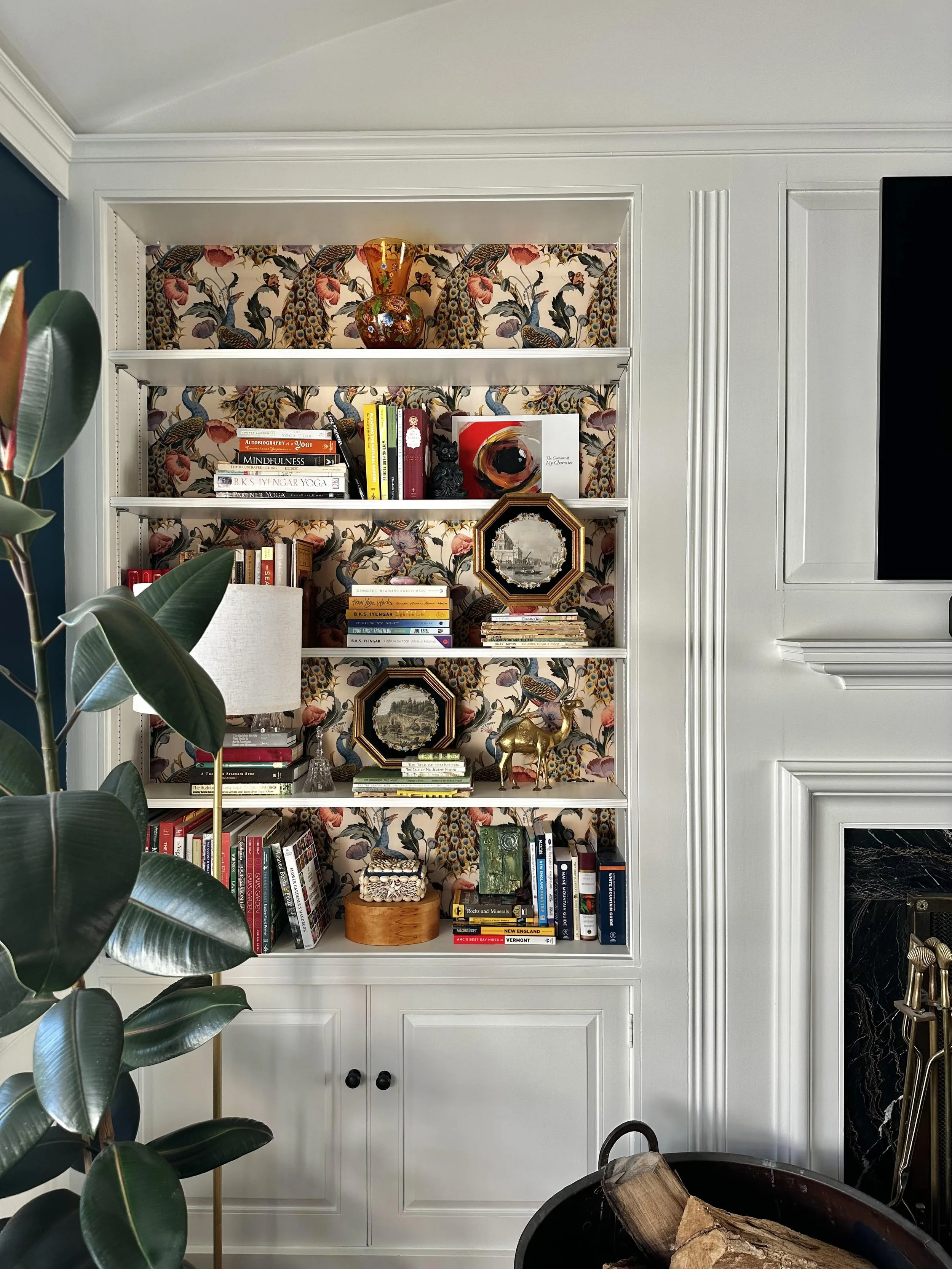 A built-in bookshelf with five shelves, decorated with books, art objects, and decorative items, against a floral wallpaper background, with a white cabinet below and fireplace to the right, and a large green plant in the foreground.