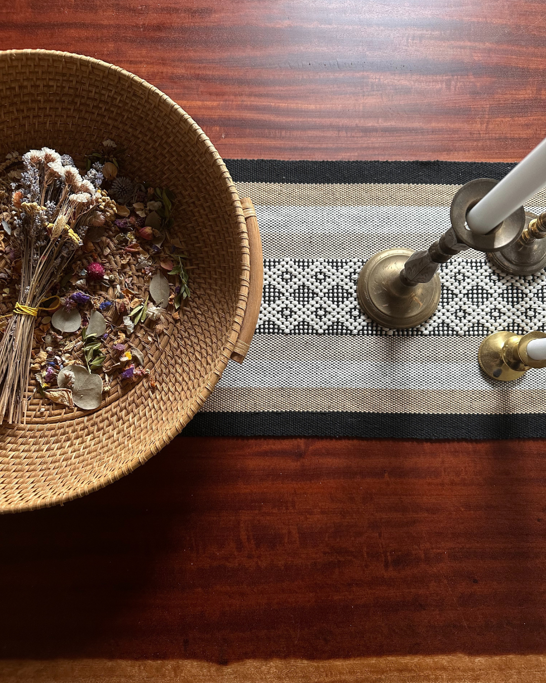 A woven basket containing dried flowers and herbs, a striped black and white table runner, brass candlesticks with candles, on a polished wooden table.