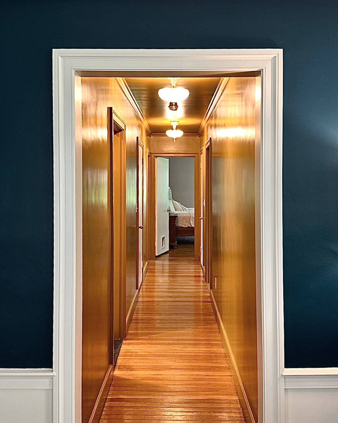 Wooden hallway with lit ceiling fixtures, leading to a bedroom with a bed and a bedside table.