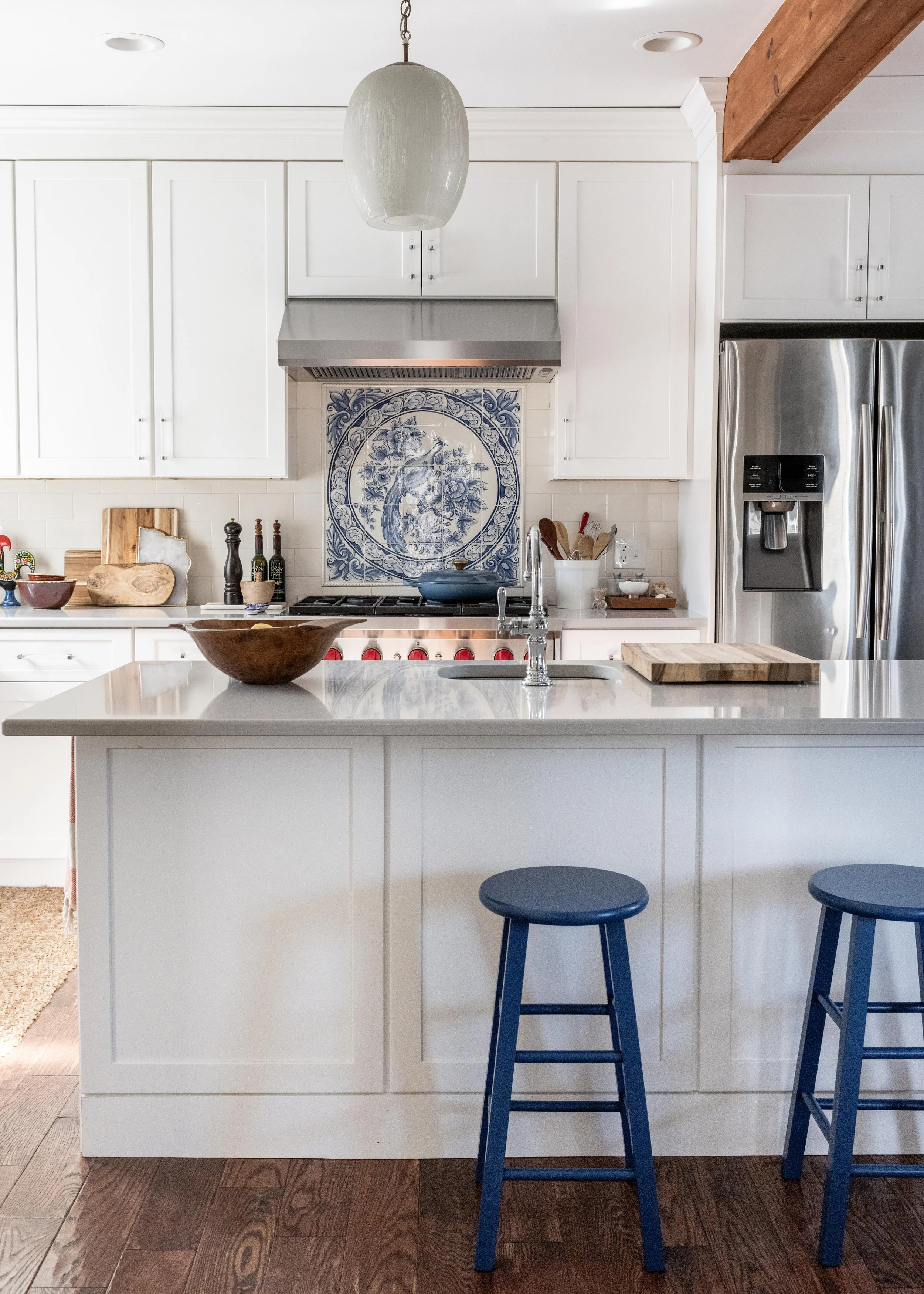 Modern white kitchen with island, blue bar stools, stainless steel refrigerator, and a blue decorative tile above the stove.