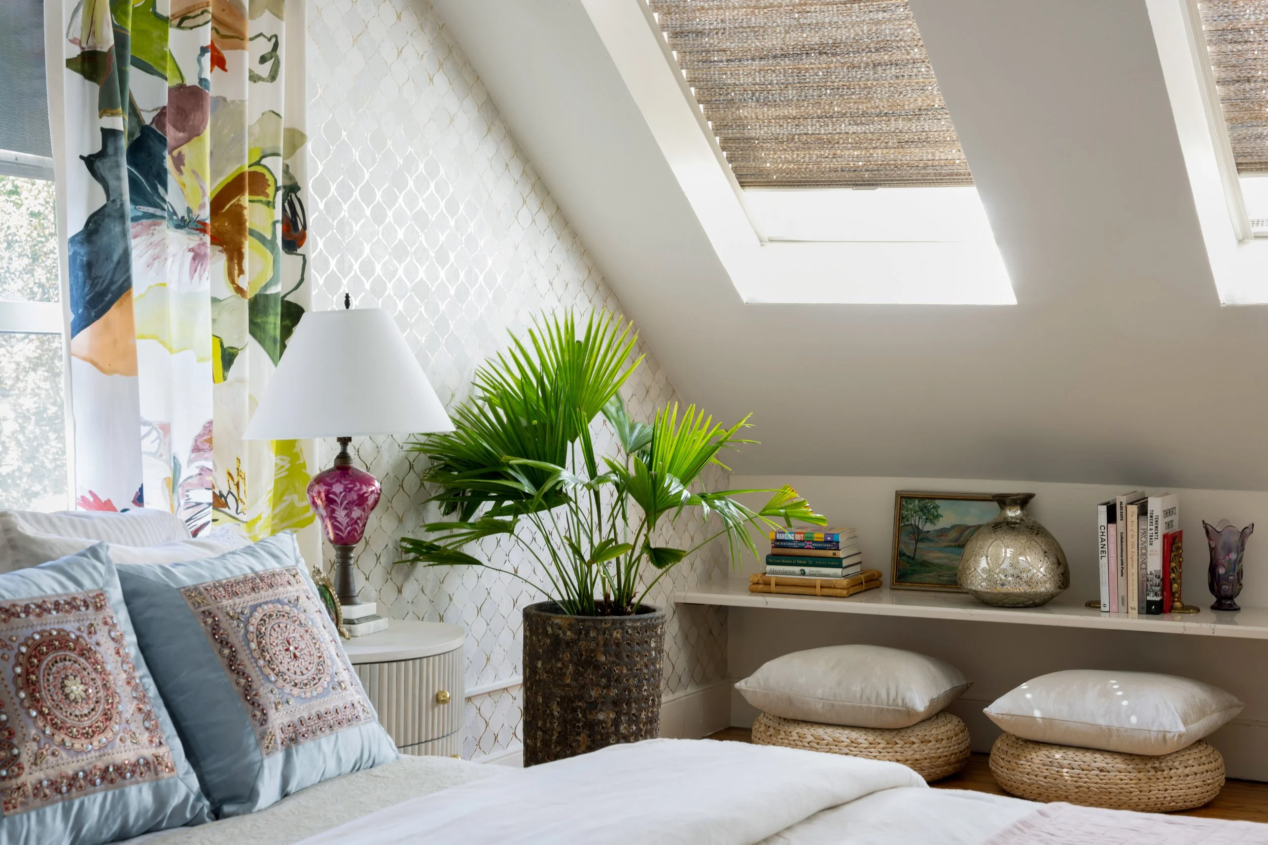 A cozy bedroom with a sloped ceiling and skylights, decorated with patterned curtains, a large green potted plant, a white nightstand with a pink lamp, pillows on the bed, a built-in white shelf with books, artwork, a silver vase, and woven cushions.