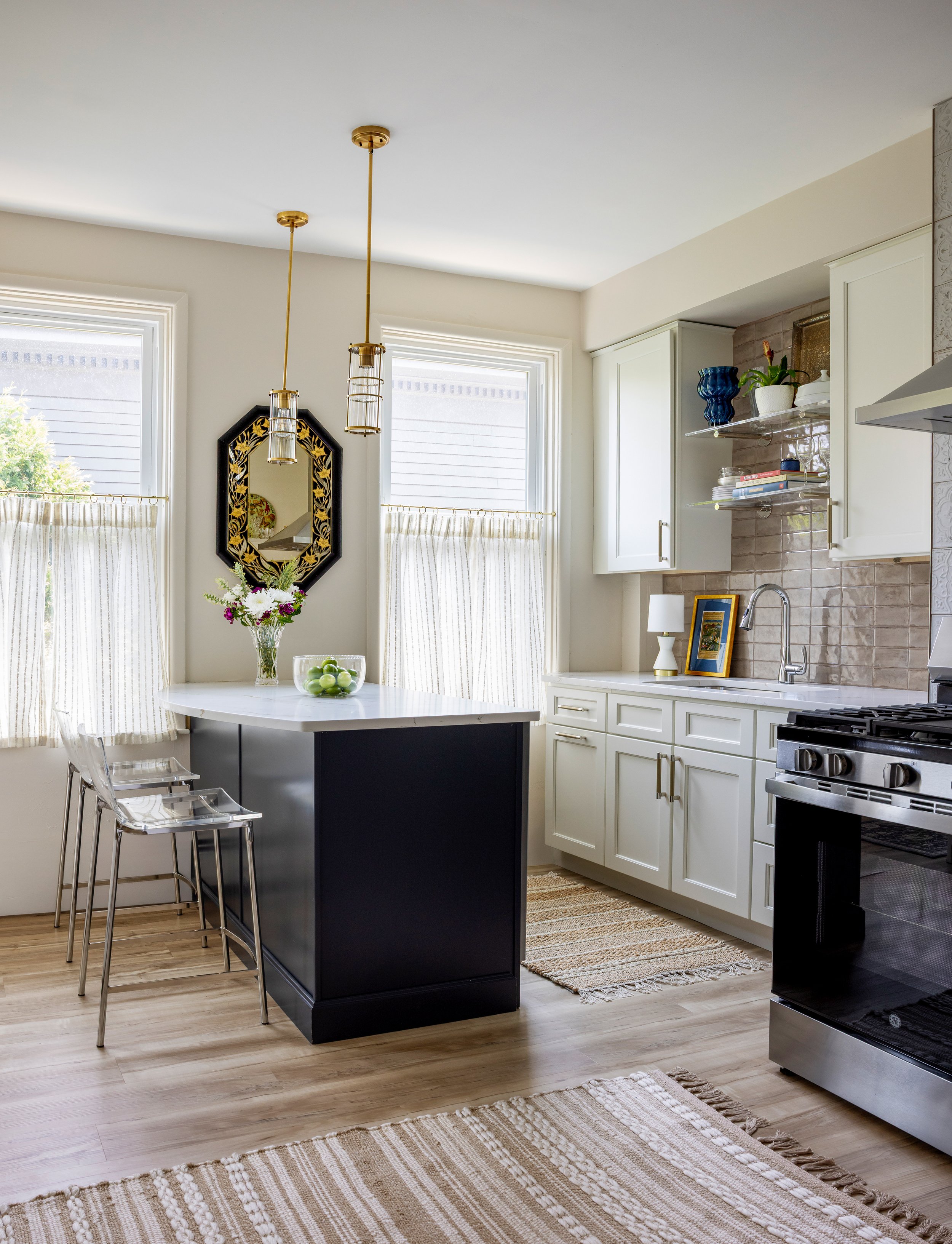 Bright kitchen with white cabinets, a black kitchen island, and two windows with striped curtains. Decor features a wall mirror, flowers, apples, and stylish pendant lights.