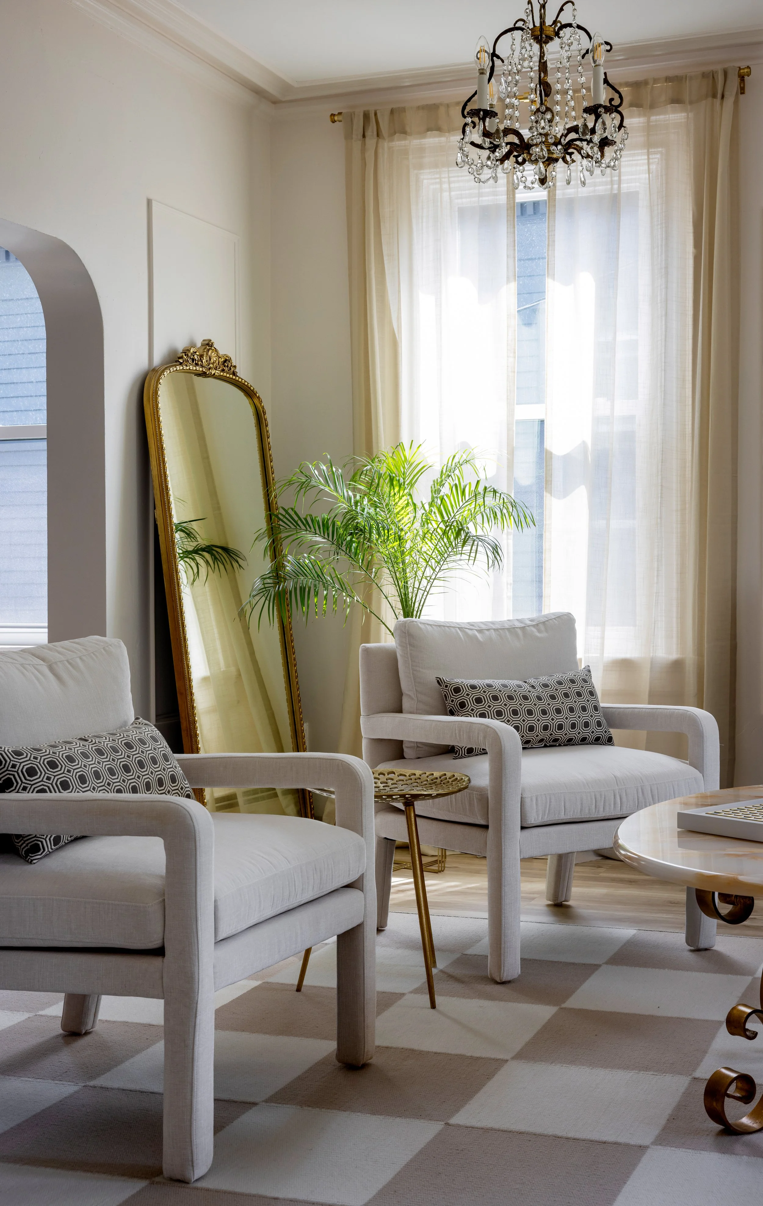 Bright living room with white armchairs, a tall mirror, a green potted plant near window with sheer curtains, and a chandelier hanging from ceiling.