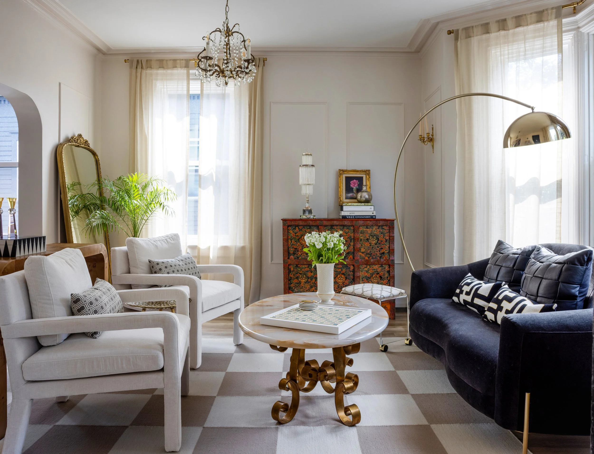 A bright living room with white sofas, a black sofa, a wooden sideboard, a large mirror, plants, and a coffee table with a vase of flowers.