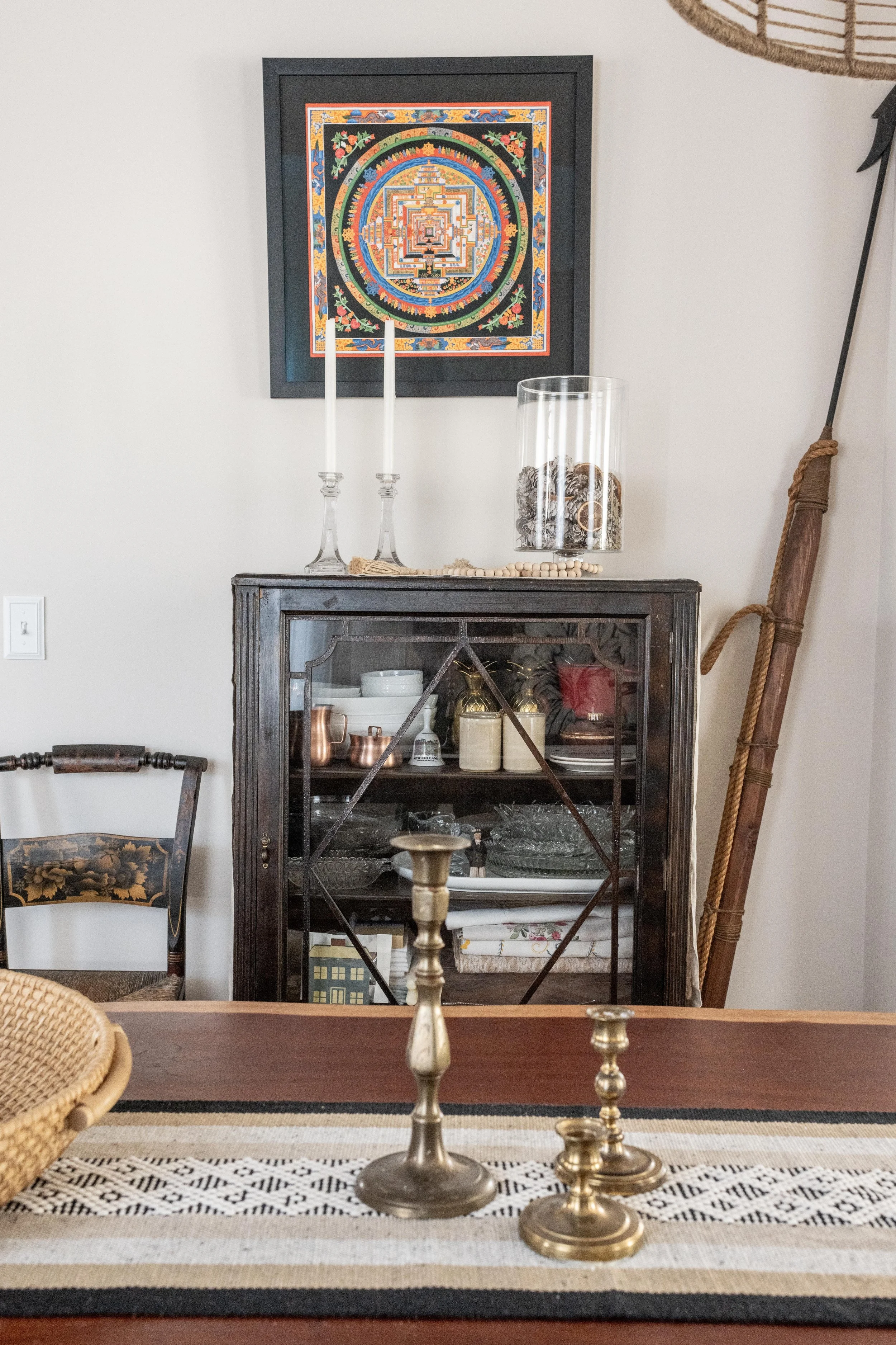 A dining room scene with a wooden table featuring three brass candle holders, a decorative runner, and a woven basket. Behind the table, there is a dark wooden glass-front cabinet containing dishes and decor items, topped with two white candles in gl
