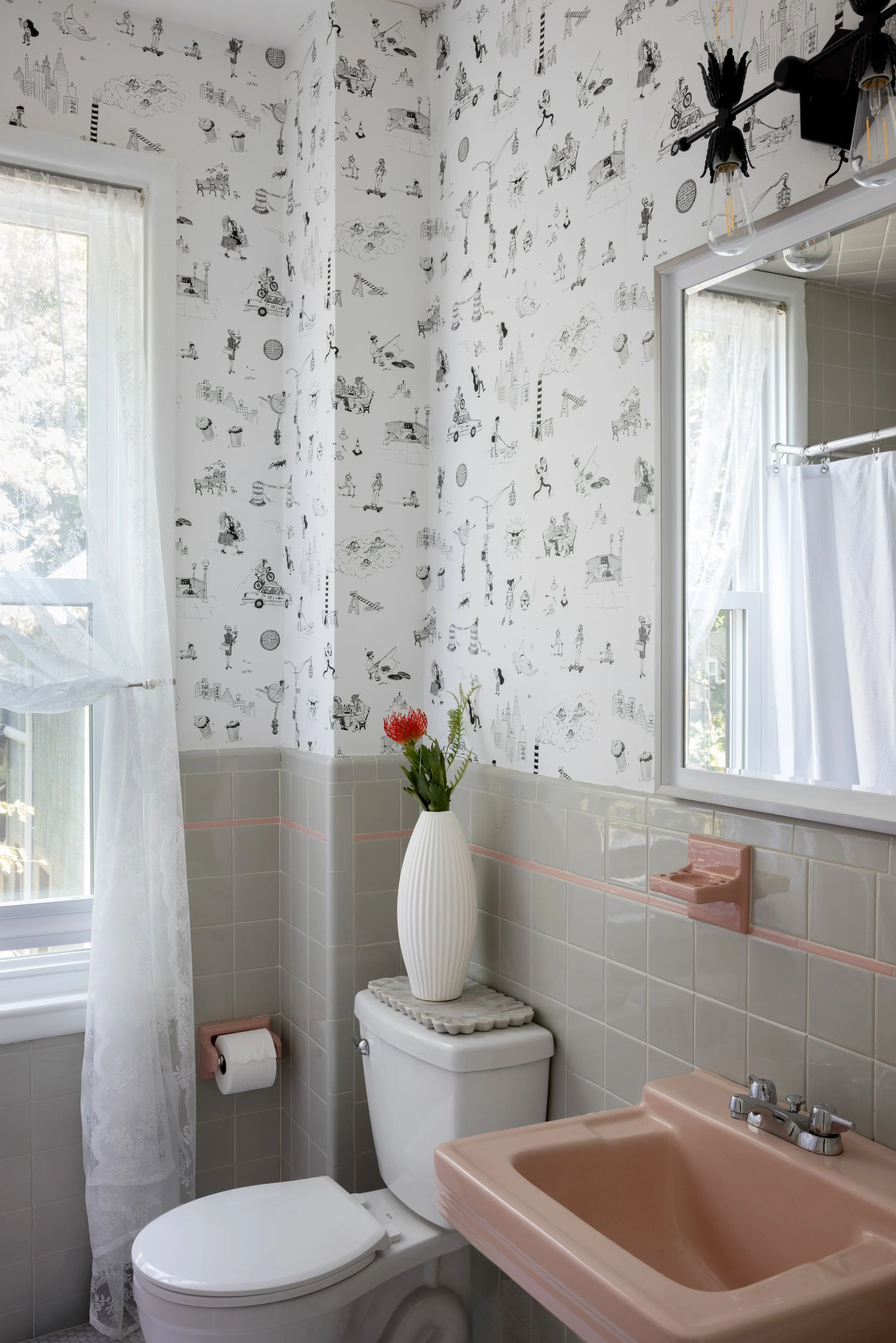 Bathroom with a pink sink and a white toilet. The walls are decorated with black-and-white sketches and illustrations. There is a window with a lace curtain, and a vase with flowers is placed on top of the toilet tank.