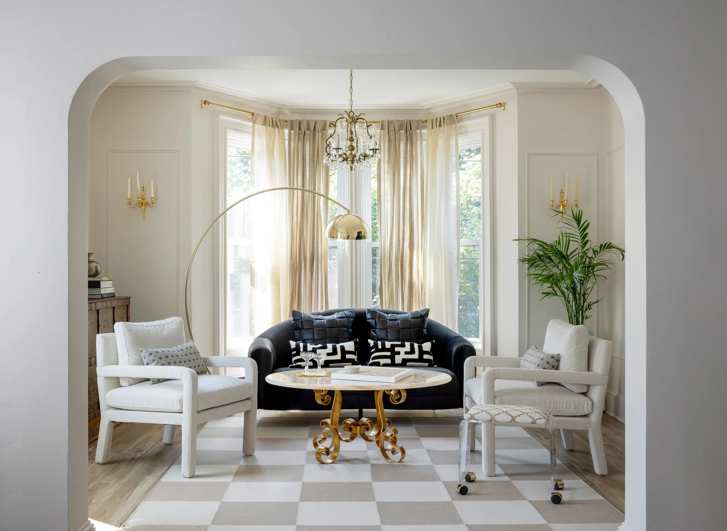 Living room with black sofa, two white armchairs, gold and marble coffee table, large windows with beige curtains, chandelier, and potted plant.