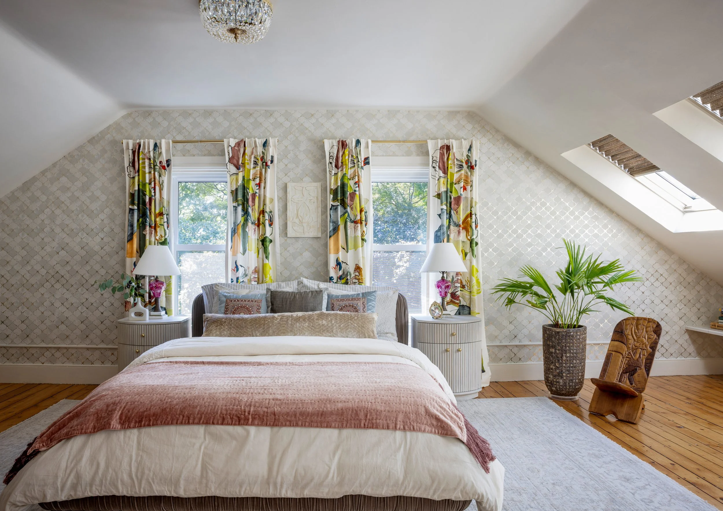 A bedroom with a sloped ceiling, two windows behind the bed, colorful floral curtains, a bed with pillows and a light pink blanket, two bedside lamps, a large potted plant, wooden flooring, a skylight window, and decorative elements.