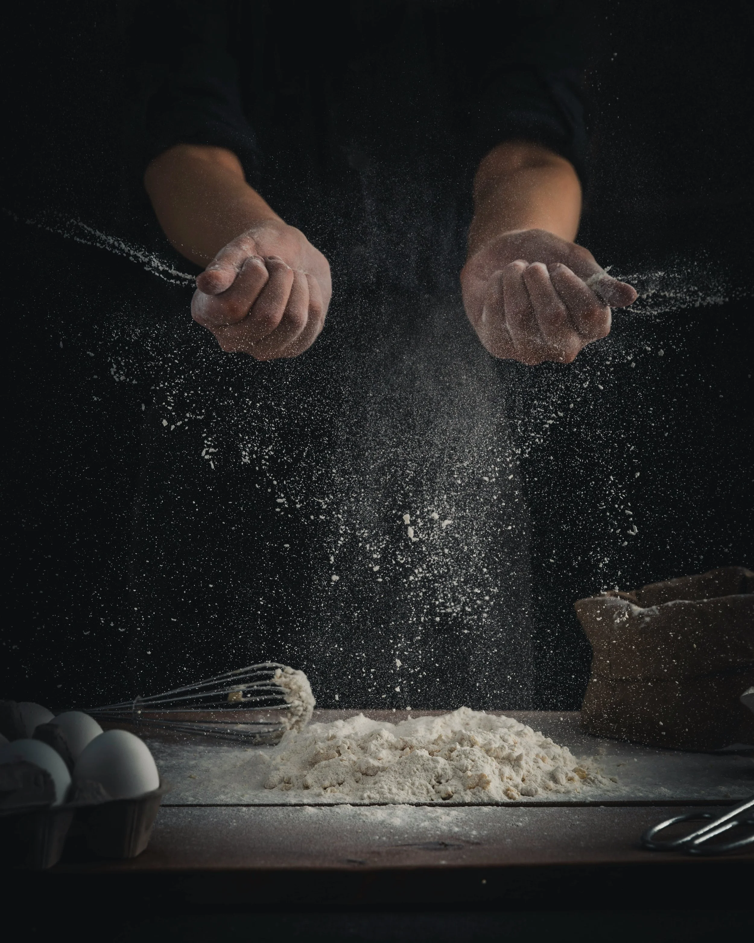 Hands in motion sifting flour over a table, with eggs in a tray, a whisk, and a bag of flour on the table.