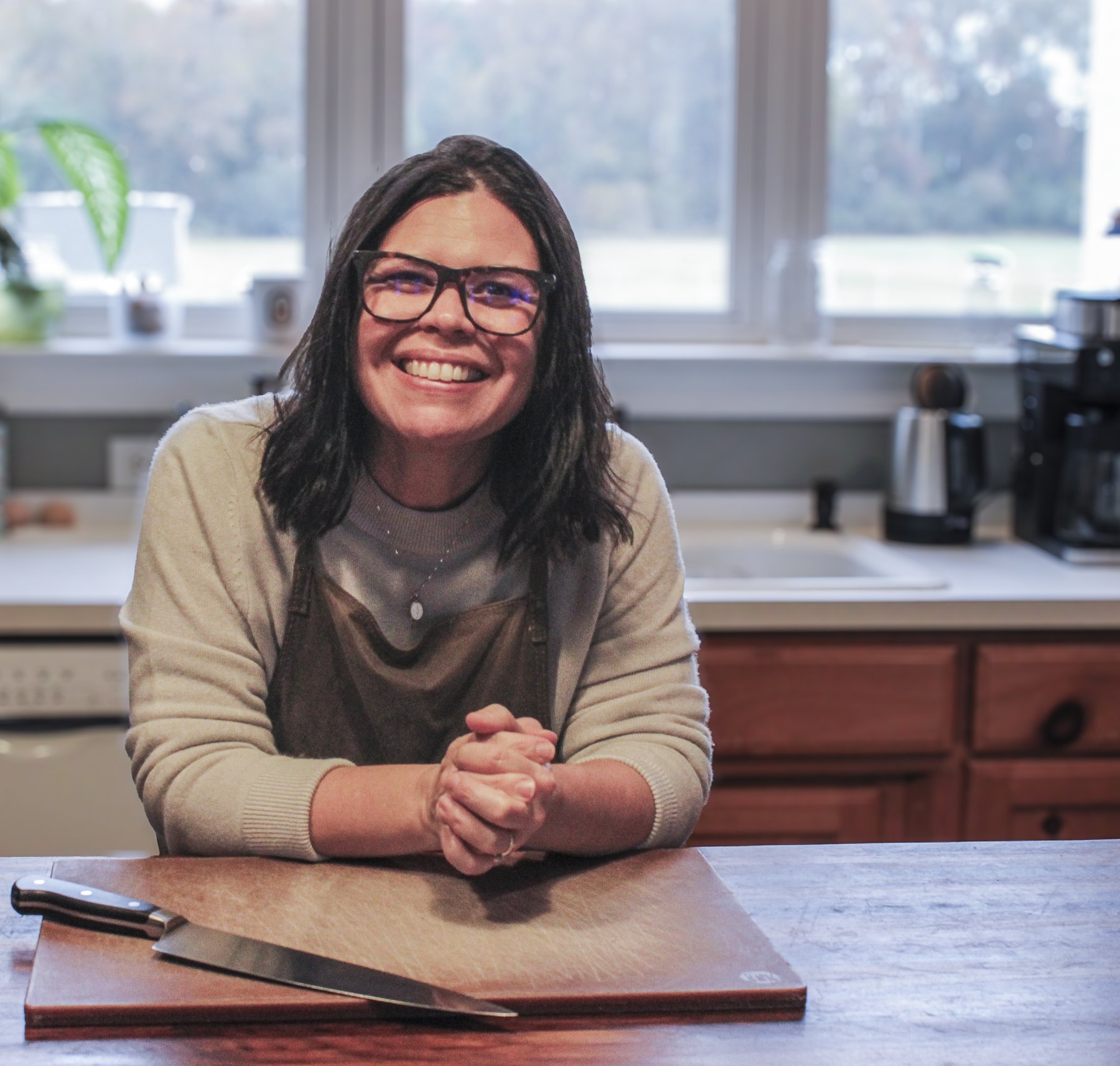 Woman with glasses smiling and sitting at a kitchen counter, with a cutting board and knife, in a well-lit kitchen with a window in the background.