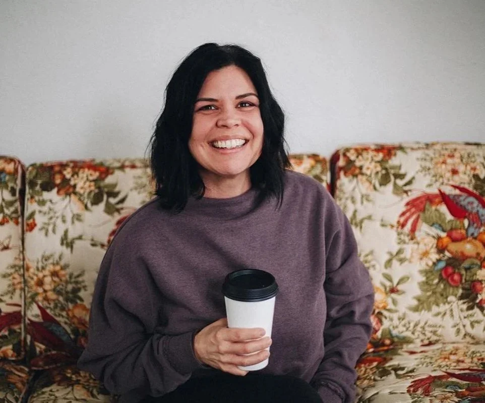 A woman with black hair smiling and holding a coffee cup, sitting on a floral-patterned couch in a cozy indoor setting.