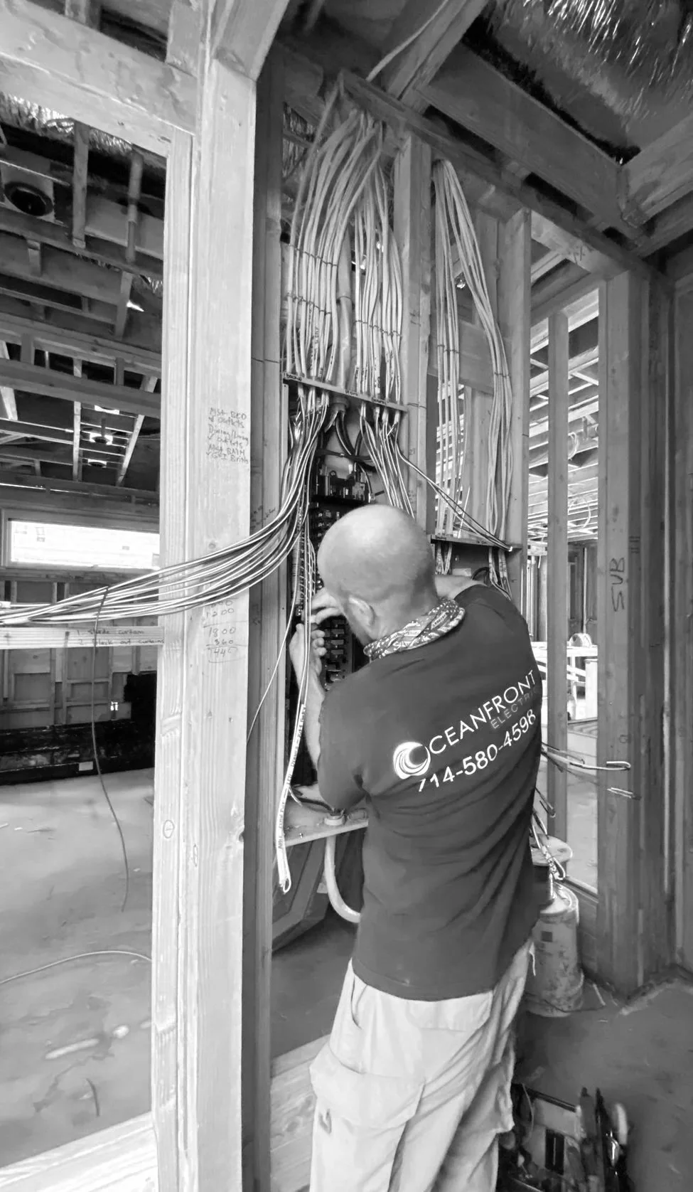 A worker with a bald head and a t-shirt that says 'OCEAN FRONT Electric' is installing or repairing electrical wiring inside a building under construction; the wooden framework and electrical cables are visible.