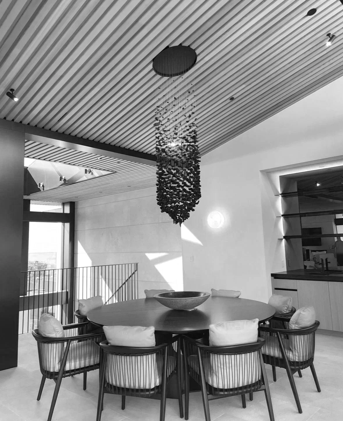 Modern dining room with round table, eight chairs with cushions, a contemporary chandelier hanging from a slatted ceiling, and a large window allowing natural light.