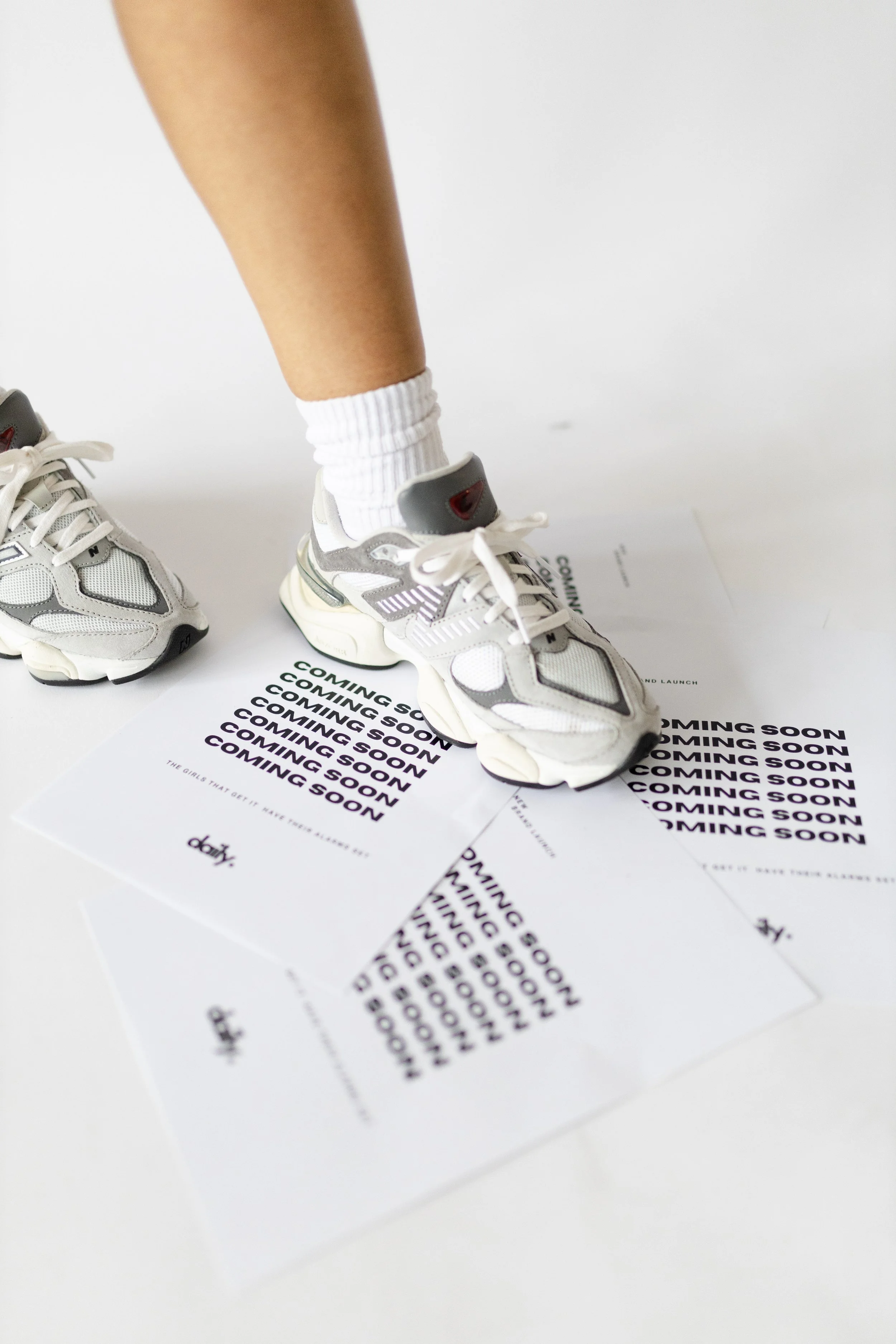 Person wearing white and gray sneakers and white socks, stepping on a white surface with 'Coming Soon' printed repeatedly. The focus is on the shoes and part of the person's leg.