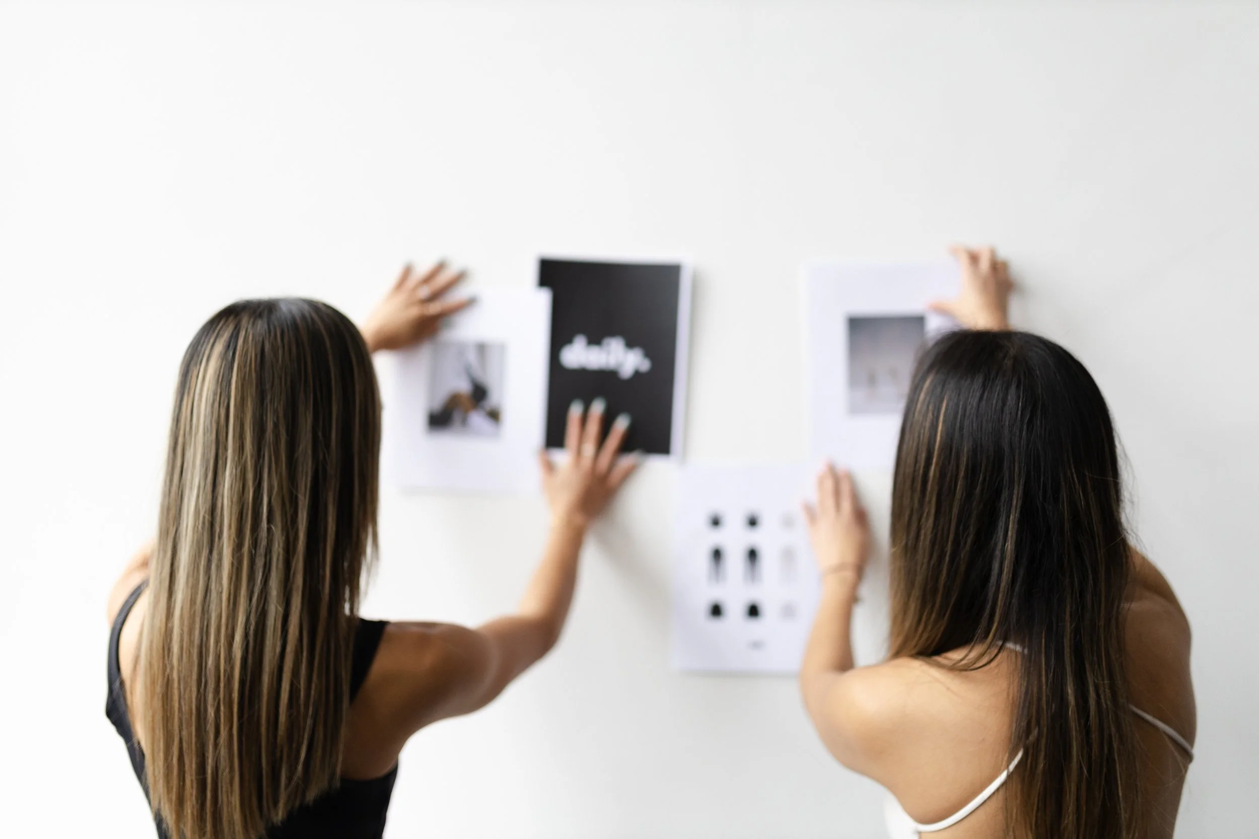 Two women with long dark hair hanging on a white wall, arranging and looking at photographs and printed materials.