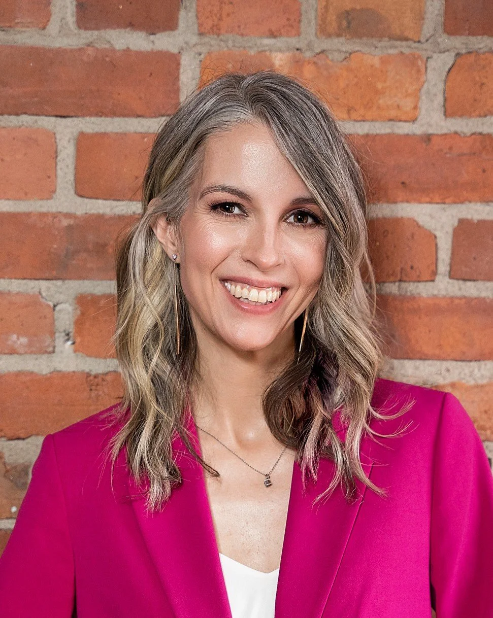 A woman with shoulder-length wavy blonde hair, wearing a bright pink blazer and earrings, smiling in front of a brick wall.