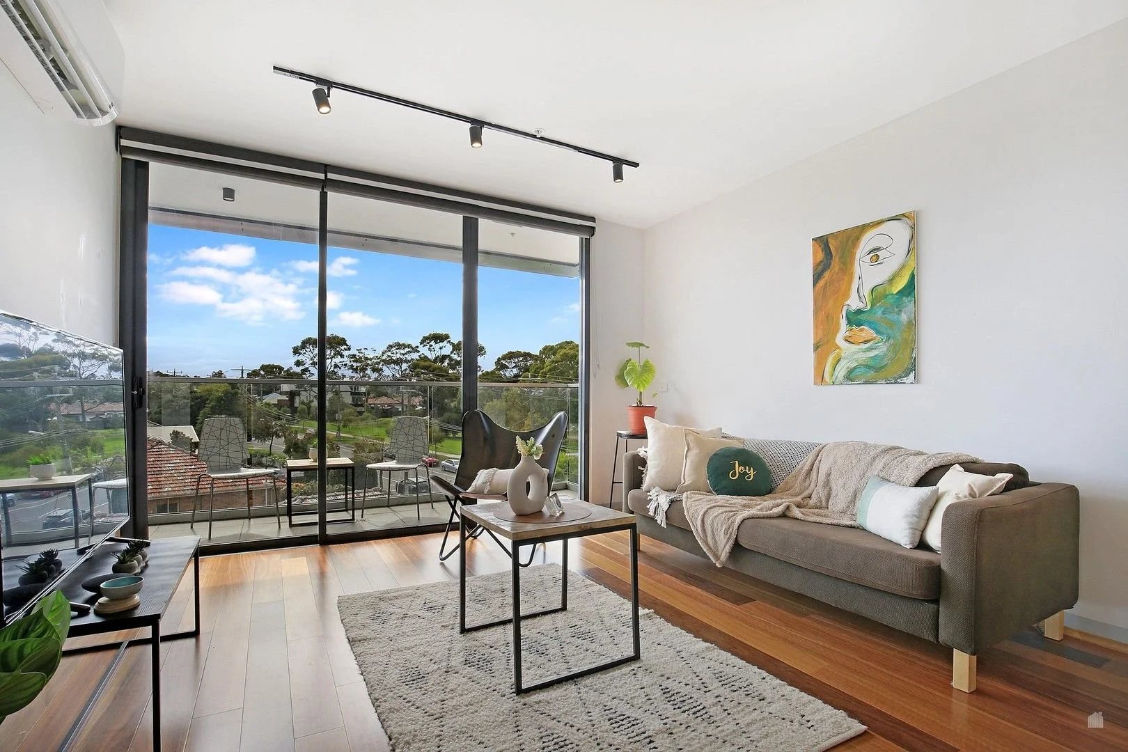 Living room with a large sliding glass door leading to a balcony, a gray sofa with pillows and a throw blanket, a coffee table with vases, and an abstract face painting on the wall.
