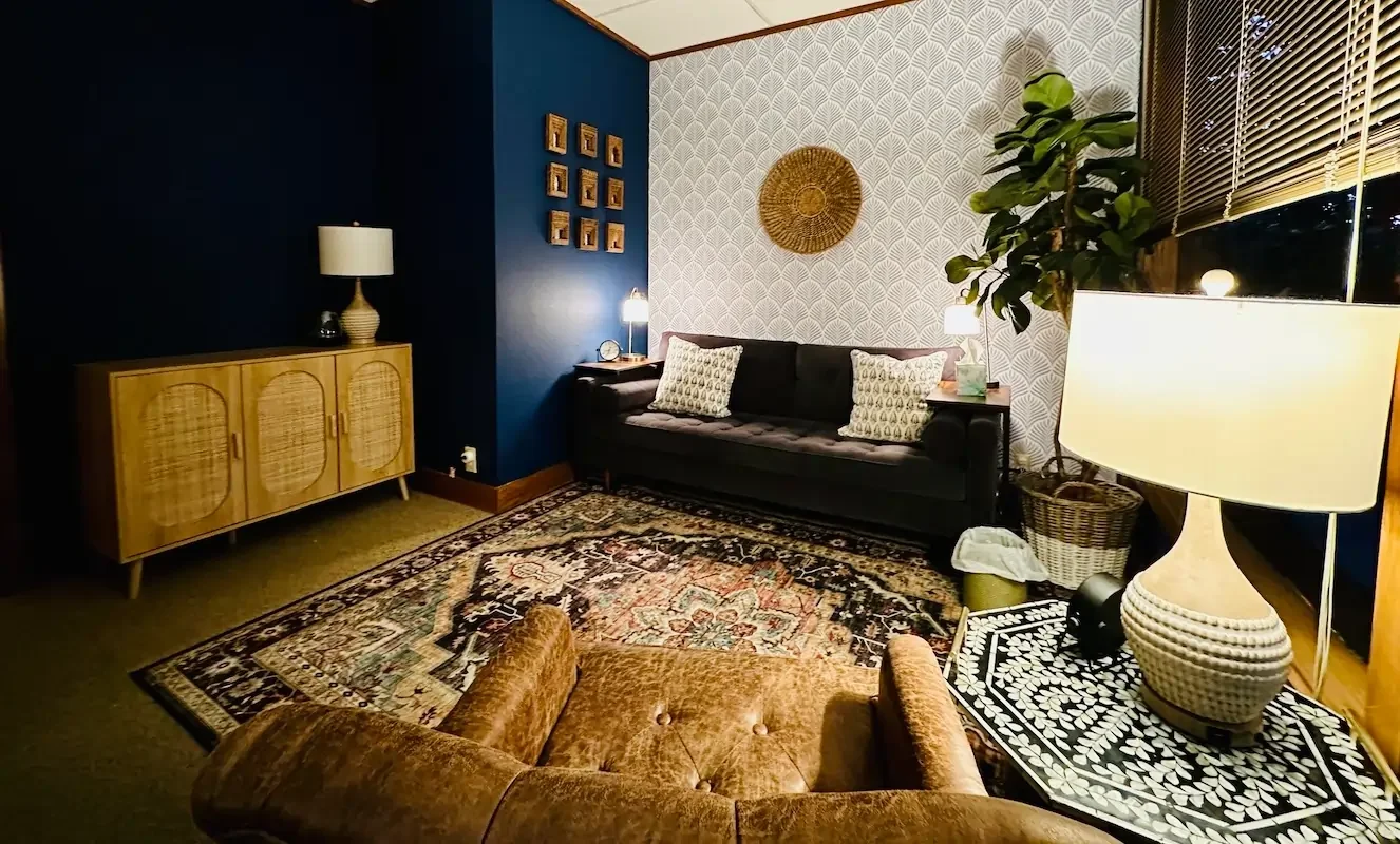 Cozy living room with dark blue and patterned wallpaper, wooden furniture, a black sofa with patterned pillows, a vintage rug, and warm lighting from table lamps and window blinds.