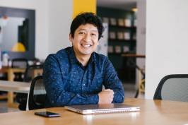 A man sitting at a desk in an office, smiling, with a laptop and smartphone in front of him.