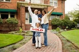 A family celebrating outside their new home with a real estate agent, holding a 'SOLD' sign.