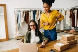 Two women in a clothing store, one sitting at a table with a laptop and the other standing, both smiling.