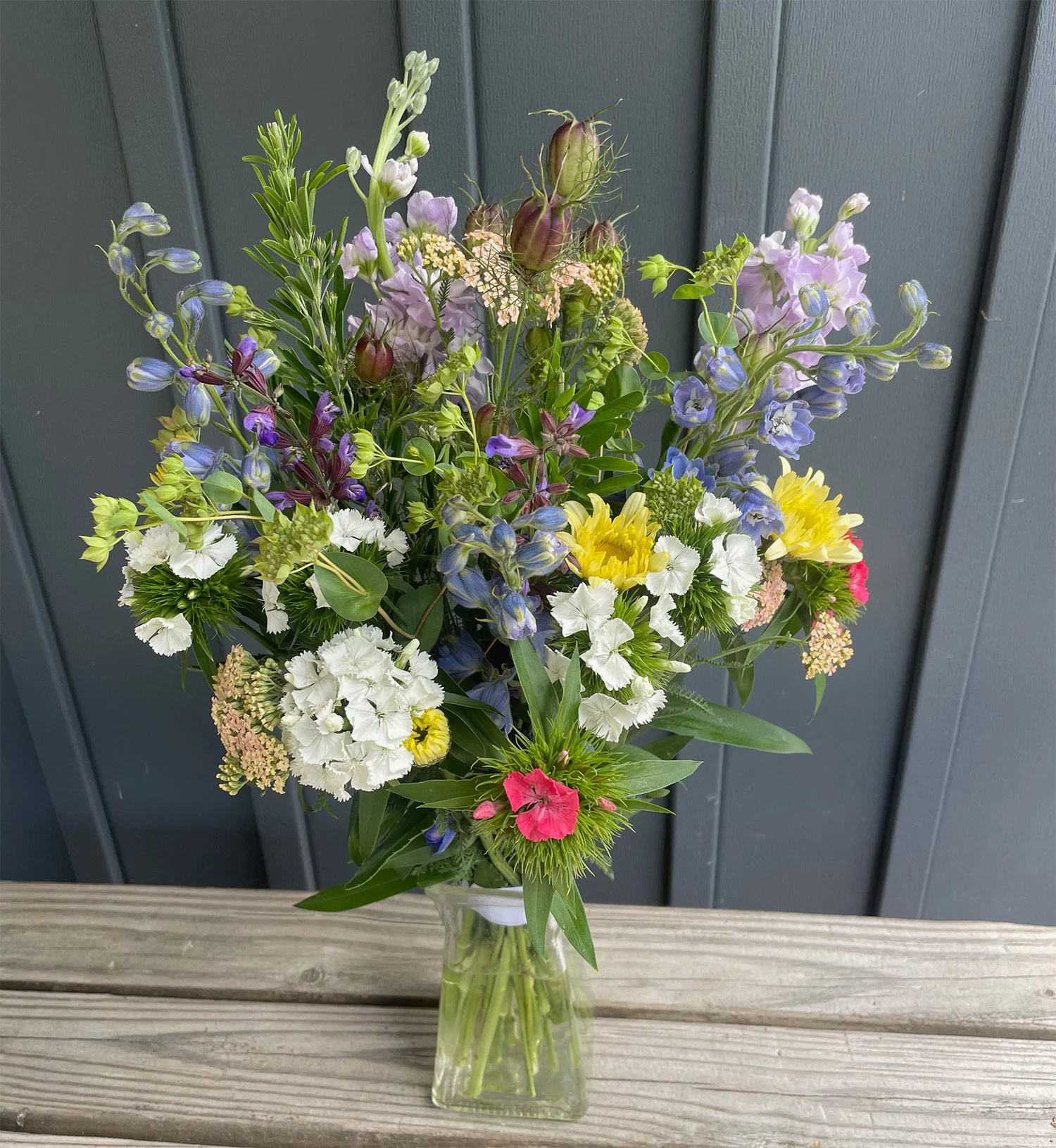 A tall glass vase filled with a colorful arrangement of fresh flowers, including white, yellow, purple, pink, and lavender blossoms, set on a wooden surface against a dark gray panel background.