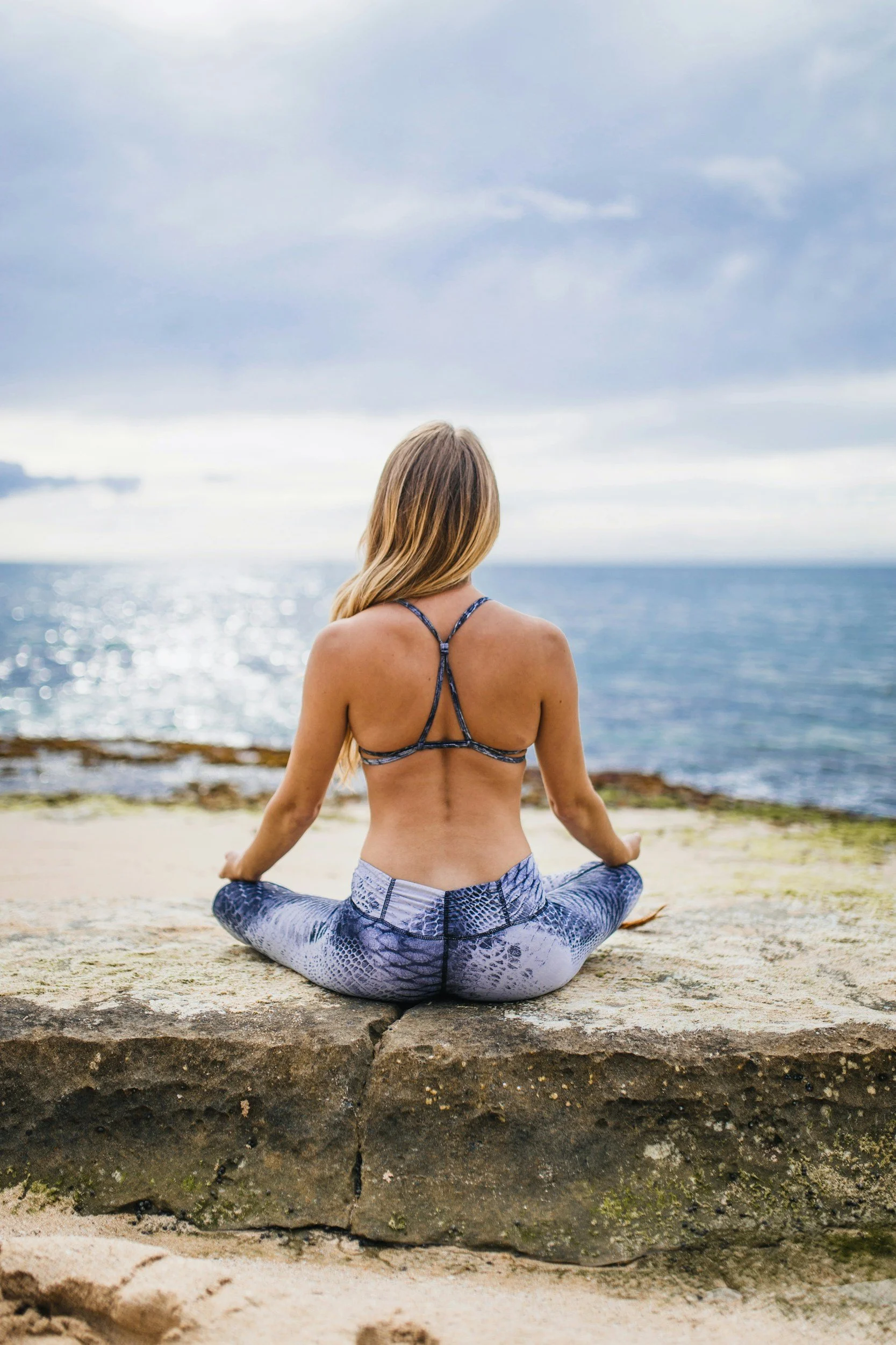 Woman meditating on a rock by the ocean in athletic wear, facing the water.