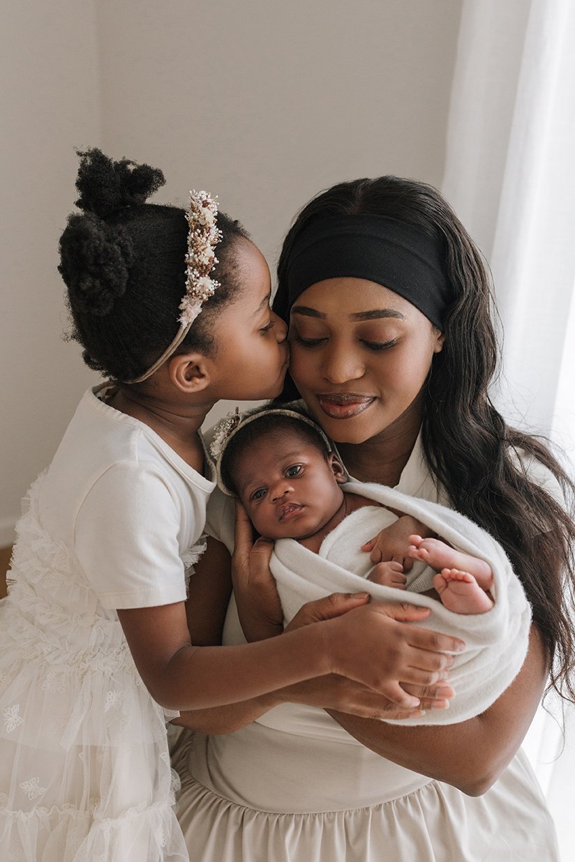 Newborn baby sleeping peacefully in mother's arms while older child kisses the mother on her head