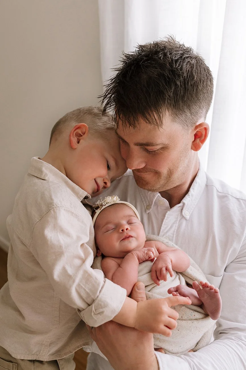 New dad, big brother and newborn baby cuddling together in a studio portrait.